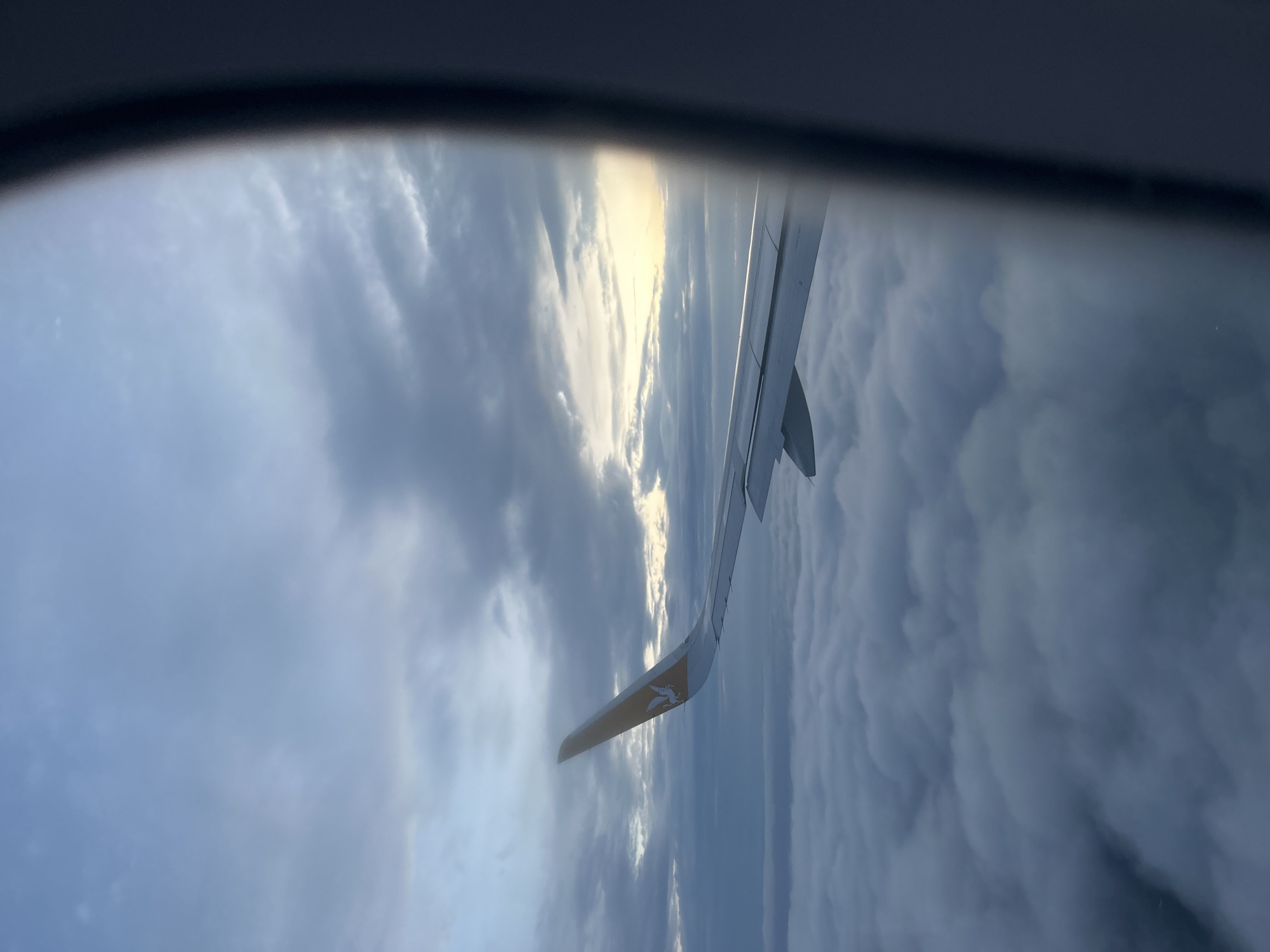 The image shows a view from an airplane window, capturing the wing of the plane against a backdrop of clouds. The wing is predominantly white with a small colored section at the wingtip bearing a logo of a stylized bird. The clouds appear varied, with darker, more textured areas interspersed with brighter patches where sunlight seems to be filtering through. The angle of the shot suggests that the wing is oriented diagonally in the frame. There is no indication of the city of Saint Petersburg, Russia. The window frame is visible at the top of the image, providing context for the aerial view.