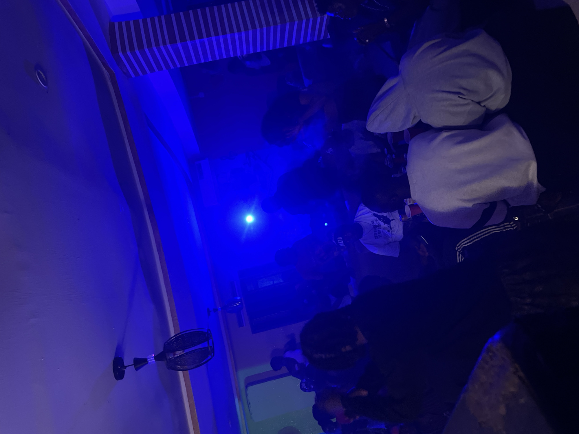 The image depicts a group of people gathered indoors, bathed in vibrant blue light. The scene appears to be a party or social gathering, likely at night given the artificial lighting. Several individuals are visible, some with their backs to the camera, and others with their faces obscured by shadows or their clothing. They are dressed in casual attire, including hoodies and t-shirts.
Notable details include a striped ceiling or backdrop in the upper portion of the image, and a small fan mounted on the wall on the left. A bright, circular light source is also visible in the center of the image, adding to the ambient lighting. The overall atmosphere seems relaxed and social. No specific time of day or weather conditions can be inferred from the image, but the indoor setting and blue lighting suggest an evening or nighttime event.
There is some visible text on a person's t-shirt in the upper right of the image. While partially obscured and difficult to read clearly due to the lighting and angle, it appears to be a graphic with text that might include "STOP THINKING START LIVING" or similar motivational phrasing. The location is noted as Durumi, Nigeria.