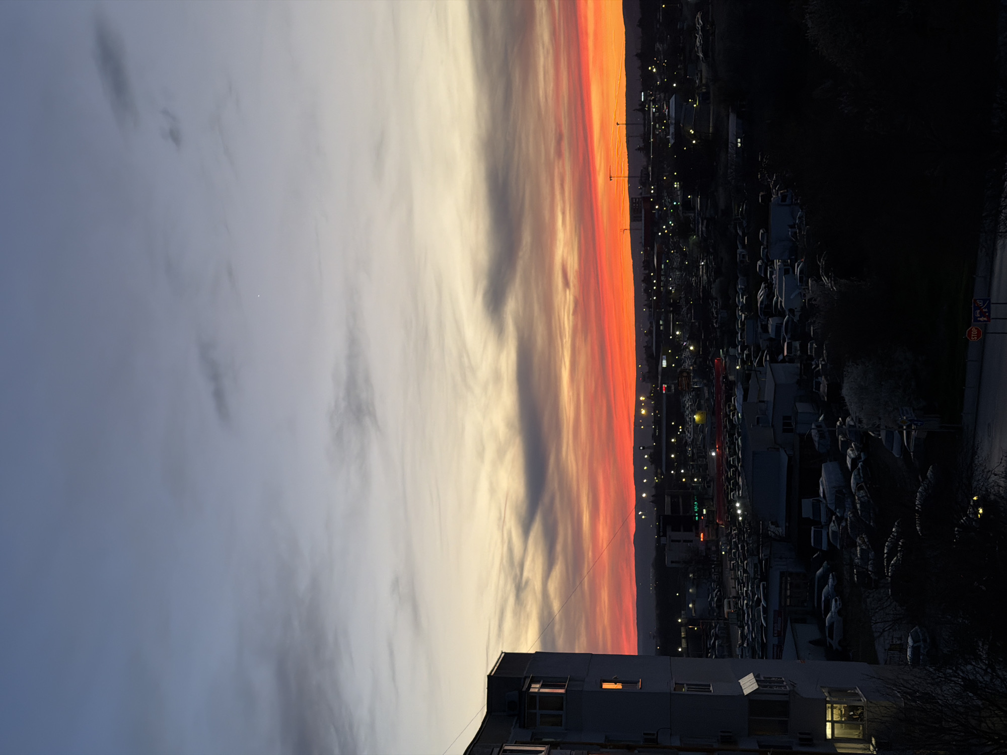 The image shows a sunset over Varna, Bulgaria, taken from an elevated position, possibly a high-rise building. The sky displays a gradient of colors, transitioning from a light gray-blue with scattered clouds on the left to vibrant orange and red hues on the right horizon. A row of buildings is visible in the lower center of the frame. Below the sunset, the image captures a dense urban landscape with numerous parked cars, streetlights, and building lights scattered throughout. The right side of the frame features silhouetted trees and dark buildings, adding contrast to the illuminated cityscape. A stop sign is visible on the lower right side of the frame.