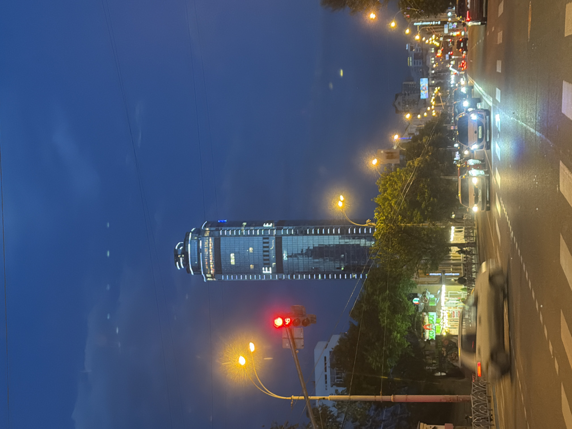 The image captures a street scene in Yekaterinburg, Russia, at dusk or night. A tall, modern skyscraper with illuminated windows dominates the background. In the foreground, a street light with a red traffic signal is visible on the left. Cars are driving on a wet road, reflecting the streetlights and building lights. Several trees and buildings line the street, some with illuminated signs. The sky is a deep blue with a few faint clouds. The overall impression is a bustling urban environment at night.