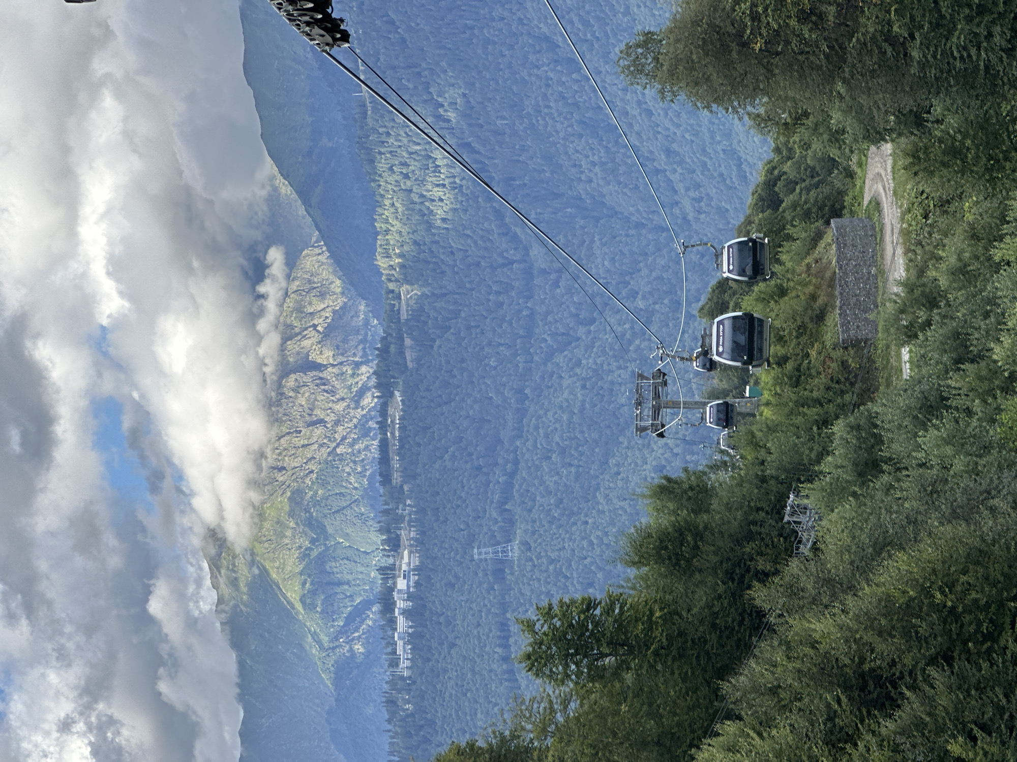 The image depicts a panoramic view of a mountain landscape with a cable car system. The cable cars, designed as enclosed cabins, are suspended from cables stretching across the frame, indicating their movement or stationary position. In the foreground, lush green trees and dense forest cover the slopes. A winding dirt road is visible amongst the trees, leading upwards.
In the mid-ground, the cable car towers are situated amidst the trees, with several cabins visible. Beyond the forested slopes, a range of majestic mountains rises, some peaks partially obscured by clouds. A large, modern building complex is visible on one of the distant mountain slopes, suggesting a resort or a research facility. A tall, lattice-like transmission tower stands on another forested hillside in the distance.
The sky is partly cloudy, with dramatic cloud formations casting shadows and highlighting portions of the mountain range. The overall scene suggests a scenic location, likely a mountain resort area.
This view is characteristic of the ski resort Rosa Khutor, located in Estosadok, Russia, a popular destination for winter sports and mountain tourism.