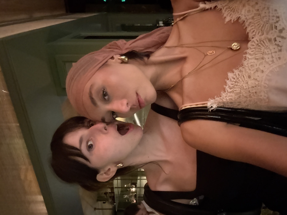 Two adult females are positioned in a close-up, likely a selfie, within a dimly lit indoor establishment. The exact location in Paris, France, is not identifiable from the image. The woman on the left has dark hair with bangs, is wearing a dark-colored square-neck top, and has gold-colored hoop earrings. Her mouth is open in an expression of surprise or playfulness, and her eyes are wide. The woman on the right, positioned slightly in front, has a sheer light pink or peach headscarf covering her hair. She is wearing multiple layered gold-colored necklaces, including one with a rectangular pendant and another with a round medallion, and gold-colored hoop earrings. Her attire consists of a light-colored (possibly white or cream) camisole with lace trim. Her expression is neutral, looking directly towards the camera. The women are in close proximity, suggesting an interaction. In the background, to the left of the woman with dark hair, a shelf with various bottles and glassware is visible, illuminated from behind. A person's head is partially visible in the far left background. The upper right portion of the image shows a section of green-paneled wall and ceiling. The overall lighting is warm and subdued, consistent with a bar or restaurant interior.