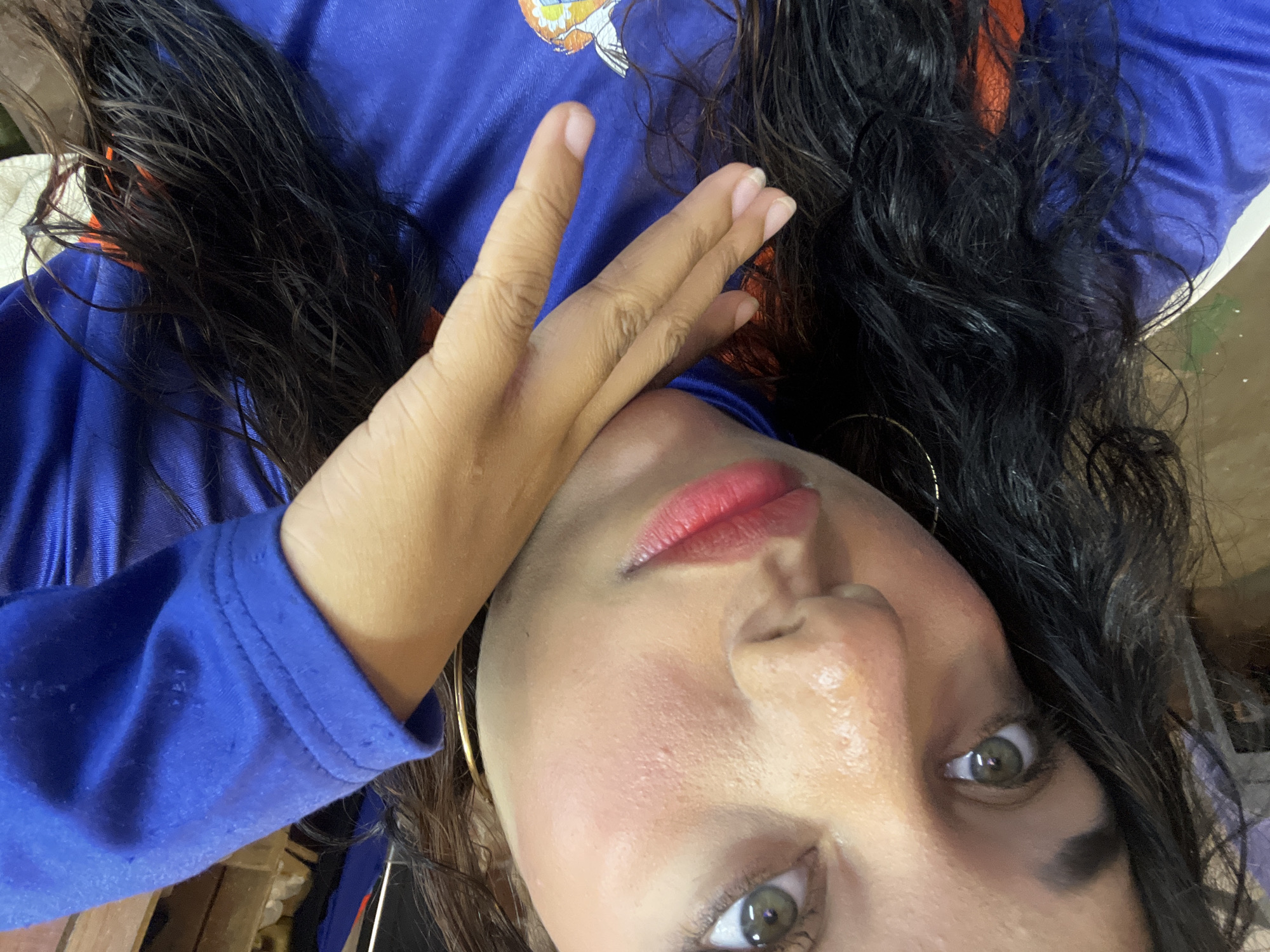 The image shows a person with dark, wavy hair and light skin wearing a blue long-sleeved shirt. The person's hand is positioned near their face, which is angled towards the camera. They have full lips with red lipstick. A gold hoop earring is visible. The person has olive/hazel eyes. The background is blurry and indistinct. There is no information in the image to suggest its location in Porlamar, Venezuela.