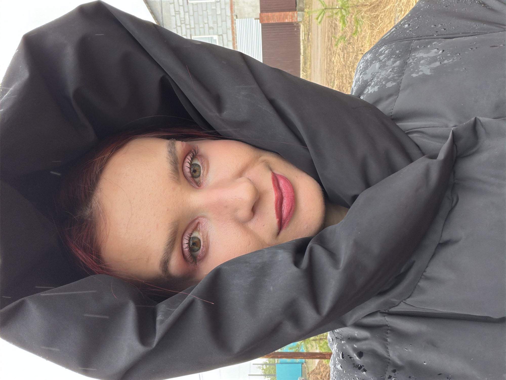 The image is a close-up portrait of a person, likely female, seen from the chest up. Her head is tilted significantly to the left within the frame, suggesting the original shot was rotated 90 degrees clockwise. She is wearing a dark, possibly black, puffy hooded jacket that prominently frames her face. Her fair skin, striking green eyes with pink eyeshadow and visible mascara, and bright pink lipstick are clearly visible. A subtle smile plays on her lips, and she looks directly towards the viewer with a calm and friendly expression. Strands of her dark reddish-brown hair are visible beneath the hood.
The jacket shows numerous water droplets, particularly on the right shoulder and sleeve, indicating recent rain or an ongoing drizzle. The background, which appears disoriented relative to the subject's natural upright posture, reveals an outdoor setting. Visible elements include a grey concrete block building, a brown corrugated metal fence, sparse green foliage, and a dirt-covered ground. This suggests a semi-rural or suburban area in Chaikovsky, Russia. The lighting is soft and diffused, characteristic of an overcast daytime. No text is visible in the image.
