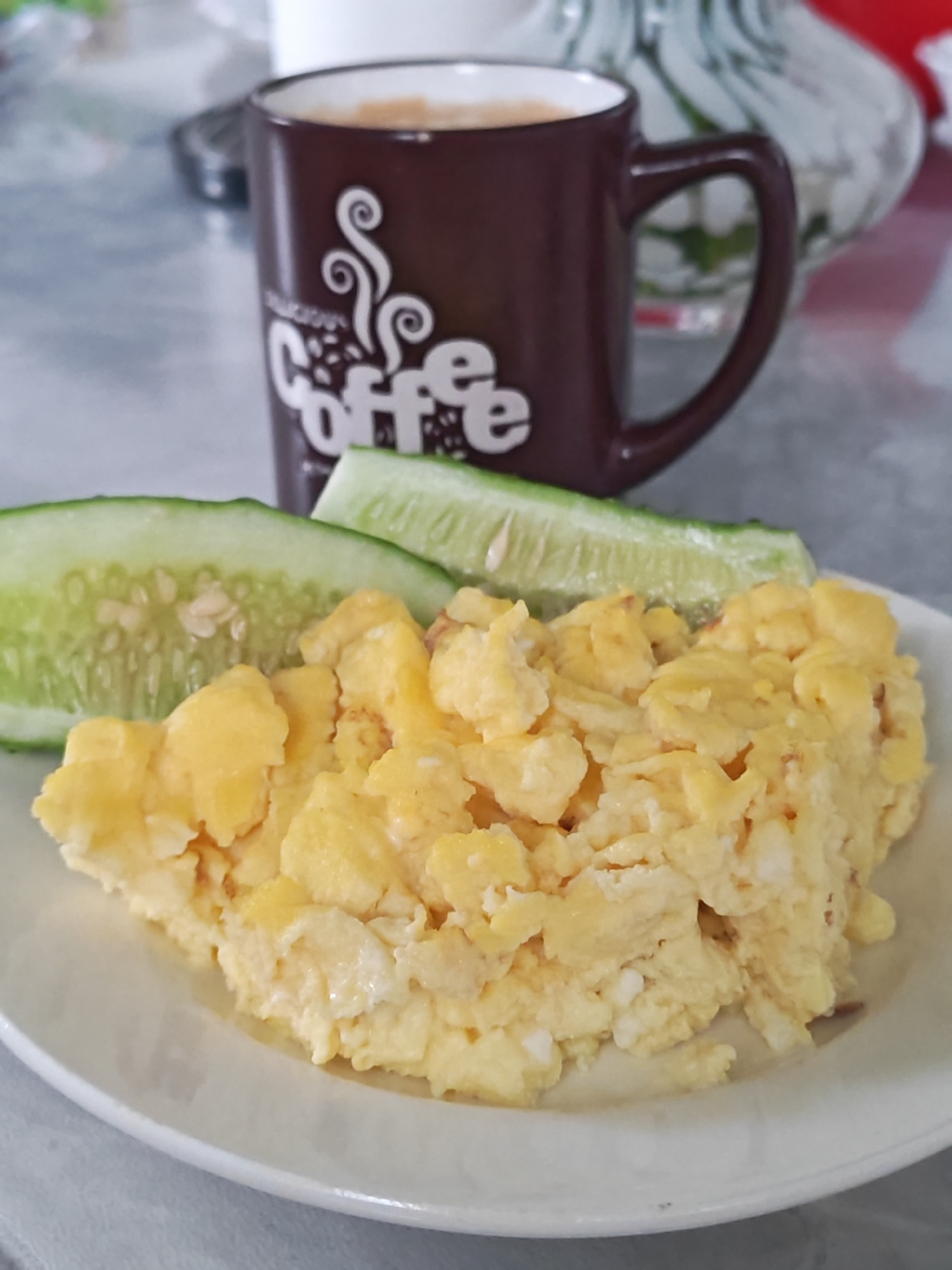 A plate of scrambled eggs with two slices of cucumber sits in the foreground. Behind the plate, a brown mug with the word "Coffee" written on it is visible. The scene is lit by natural light, and the background is slightly blurred. The exact location is not discernible from the image, and there is no evidence to suggest it is in Rybolovo, Russia. No people are visible.