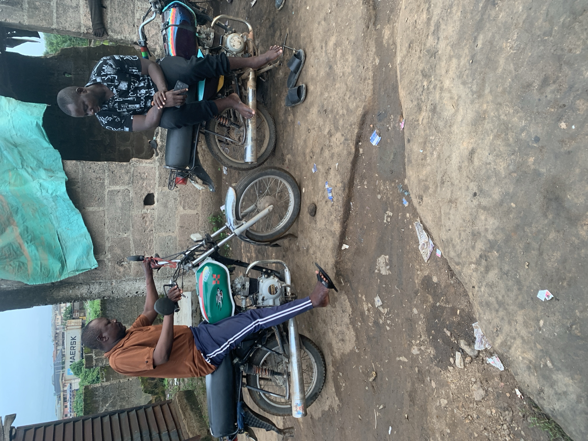 Two men are sitting on motorcycles in an outdoor area. The man on the left, wearing a brown shirt and blue pants, is on a green and white motorcycle. He is looking down at the motorcycle. The man on the right, wearing a black and white patterned shirt and black pants, is on a blue and black motorcycle. He is holding a smartphone in his right hand and looking down. Both men are barefoot. The ground is dirt and appears wet in places. There are several pieces of trash scattered on the ground. In the background, there is a brick building with a blue tarp covering part of it. Further in the background, there are buildings and a shipping container with the word "MAERSK" on it. This scene is consistent with an outdoor setting in Shagamu, Nigeria, likely an area where motorcycles are commonly used or repaired.