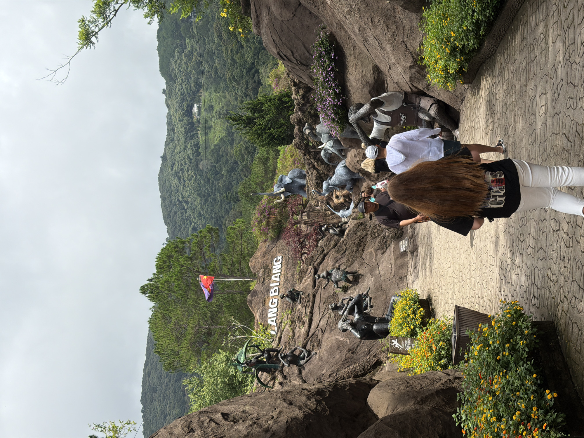 The image depicts the Lang Biang landmark in Da Lat, Vietnam. A paved pathway leads through a rocky, landscaped area adorned with various statues, including human figures and elephants. In the background, a lush, green mountainside is visible under a cloudy sky. Several people are walking on the pathway. A woman with long brown hair in a black t-shirt and white pants is in the foreground, facing away from the camera. Other individuals are seen further up the path, some wearing hats and casual clothing. The "LANG BIANG" text is displayed prominently on the rocks, accompanied by a Vietnamese flag. The environment features potted plants with yellow and orange flowers, as well as larger bushes with purple flowers.