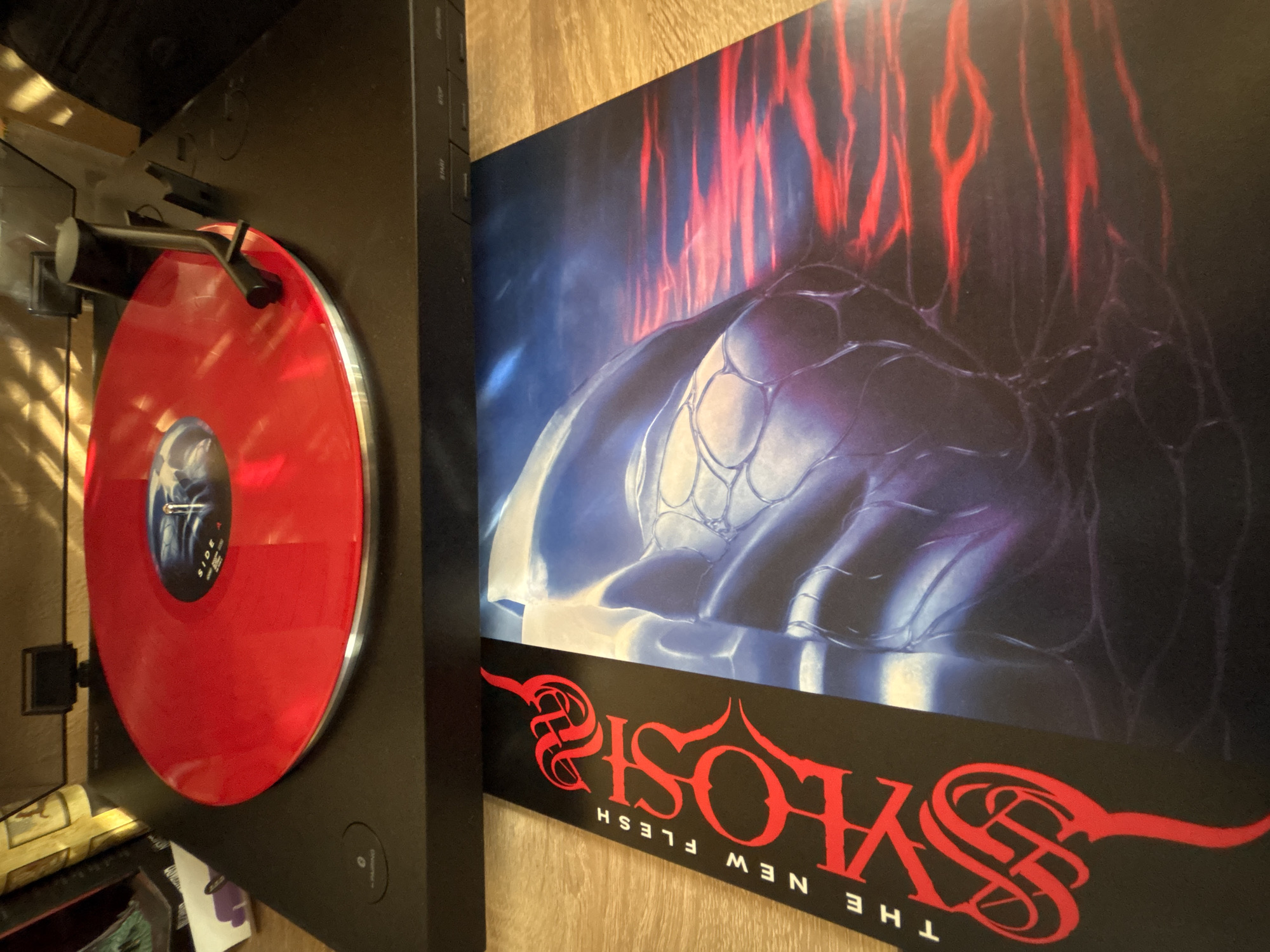 The image shows a turntable playing a red vinyl record. To the right of the turntable is a poster of the album "Syosis - The New Flesh". The environment is a room with wooden paneling. The location in Sofia, Bulgaria is not discernible from the image.