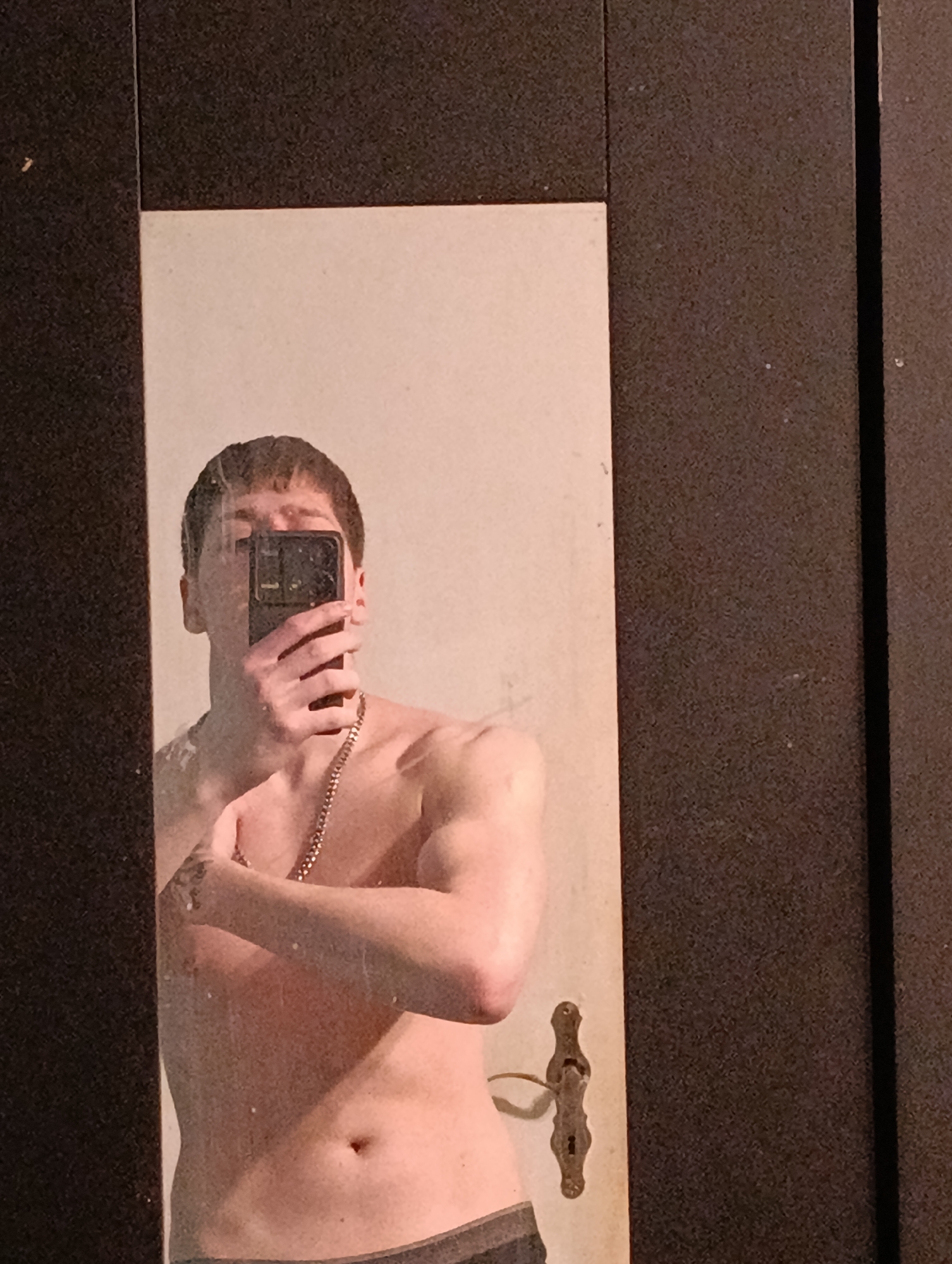 A shirtless man takes a mirror selfie in what appears to be an old door, likely a bathroom door, due to the presence of a door handle and keyhole in the mirror's reflection. He is holding a black smartphone up to his face, obscuring his eyes and nose. He has short, dark hair and is wearing a gold chain around his neck. His left arm is across his chest, revealing a visible tattoo on his forearm. The surrounding door frame is dark brown, possibly wood, and appears worn. There is no readily apparent information in the image to link it to the city of Emmerich on the Rhine, Germany.