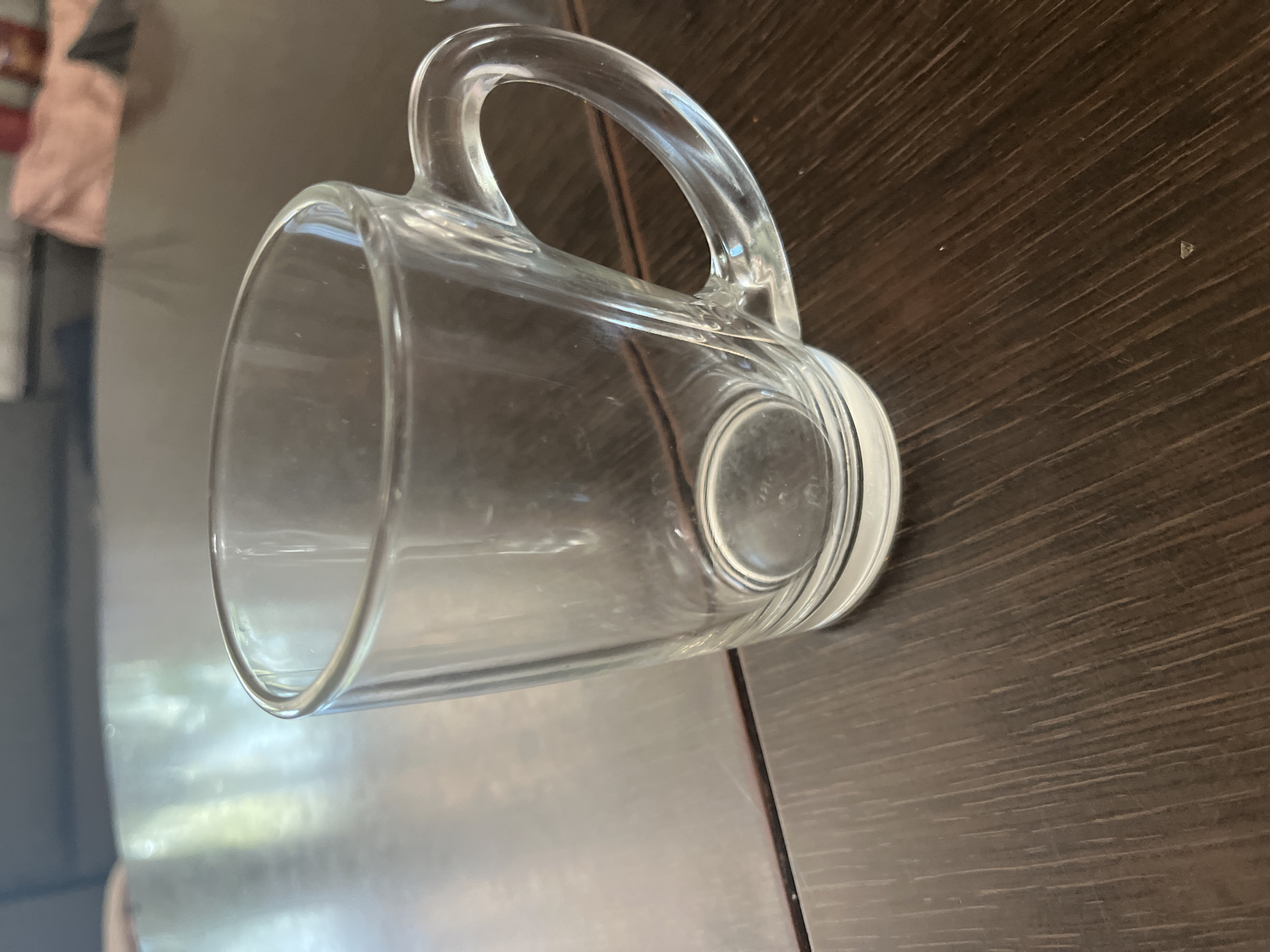 The image shows a close-up, slightly angled view of a clear glass mug resting on a dark brown wooden surface. The mug has a curved handle. The lighting suggests an indoor setting, possibly with natural light reflecting on the glass, creating subtle highlights and shadows. The background is blurred, but hints of a reflective surface and some fabric or clothing in muted colors are visible to the left, suggesting the mug is on a table or counter. There is no text visible in the image. The scene is static and peaceful, with no active event taking place.