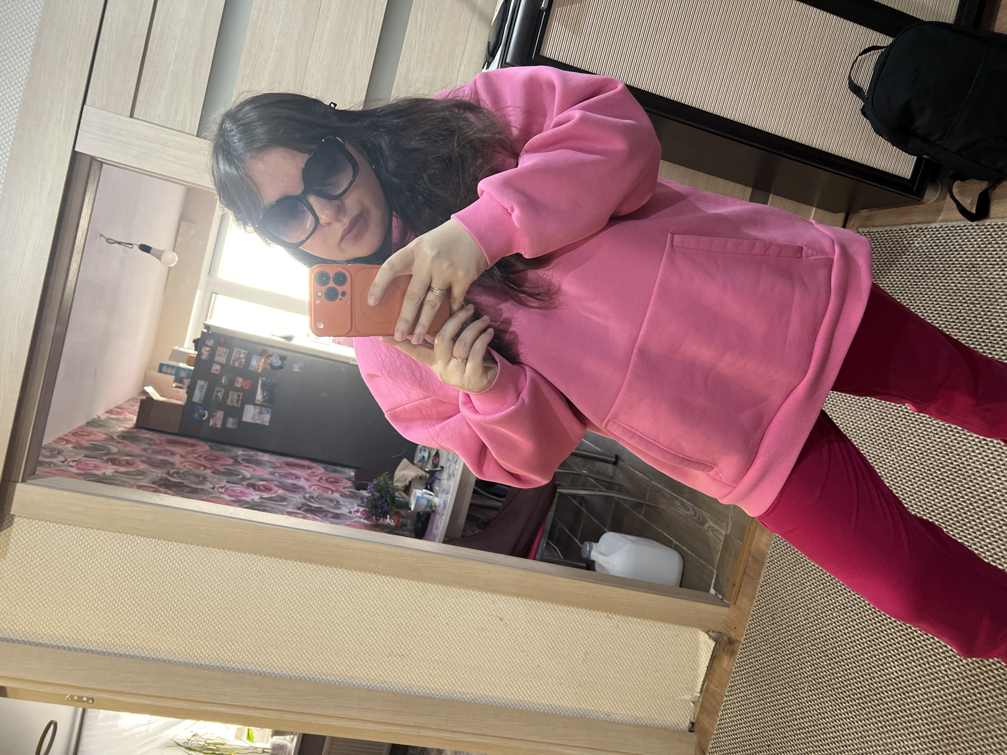 A woman wearing large sunglasses, a pink hoodie, and red pants is taking a selfie in a mirror. The reflection reveals a kitchen with floral wallpaper and various objects on the counter and refrigerator, suggesting a domestic setting. A black backpack sits on a woven rug. The exact location within Noviy, Russia, cannot be determined from the image.