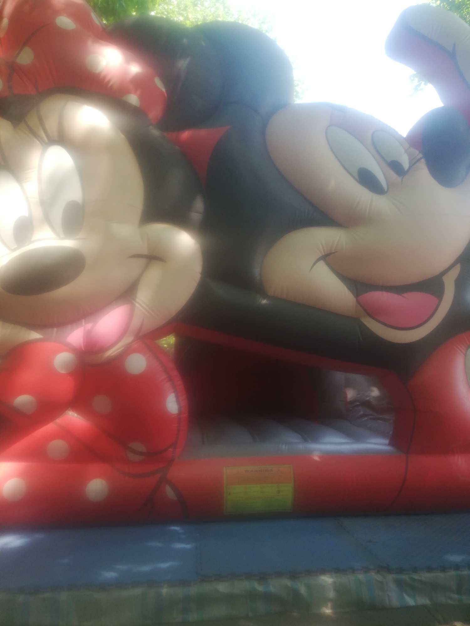 The image shows a close-up, low-angle shot of a large inflatable bounce house featuring characters from Mickey Mouse. On the left is Minnie Mouse, depicted from the chest up, with her signature red and white polka-dot bow prominently displayed. Her large, round eyes and smiling face are visible. To the right of Minnie is Mickey Mouse, also shown from the chest up. His large, round eyes and wide, open-mouthed smile with a pink tongue are characteristic. Both characters are inflated and appear to be part of a larger structure, likely a bounce house or inflatable play area. The background consists of blurred greenery and bright, overexposed sky, suggesting an outdoor setting. A red inflatable portion with a yellow warning label is visible at the bottom center of the image, indicating it's part of the bounce house structure. The image does not provide any specific visual cues to identify its location in Yerevan, Armenia.
