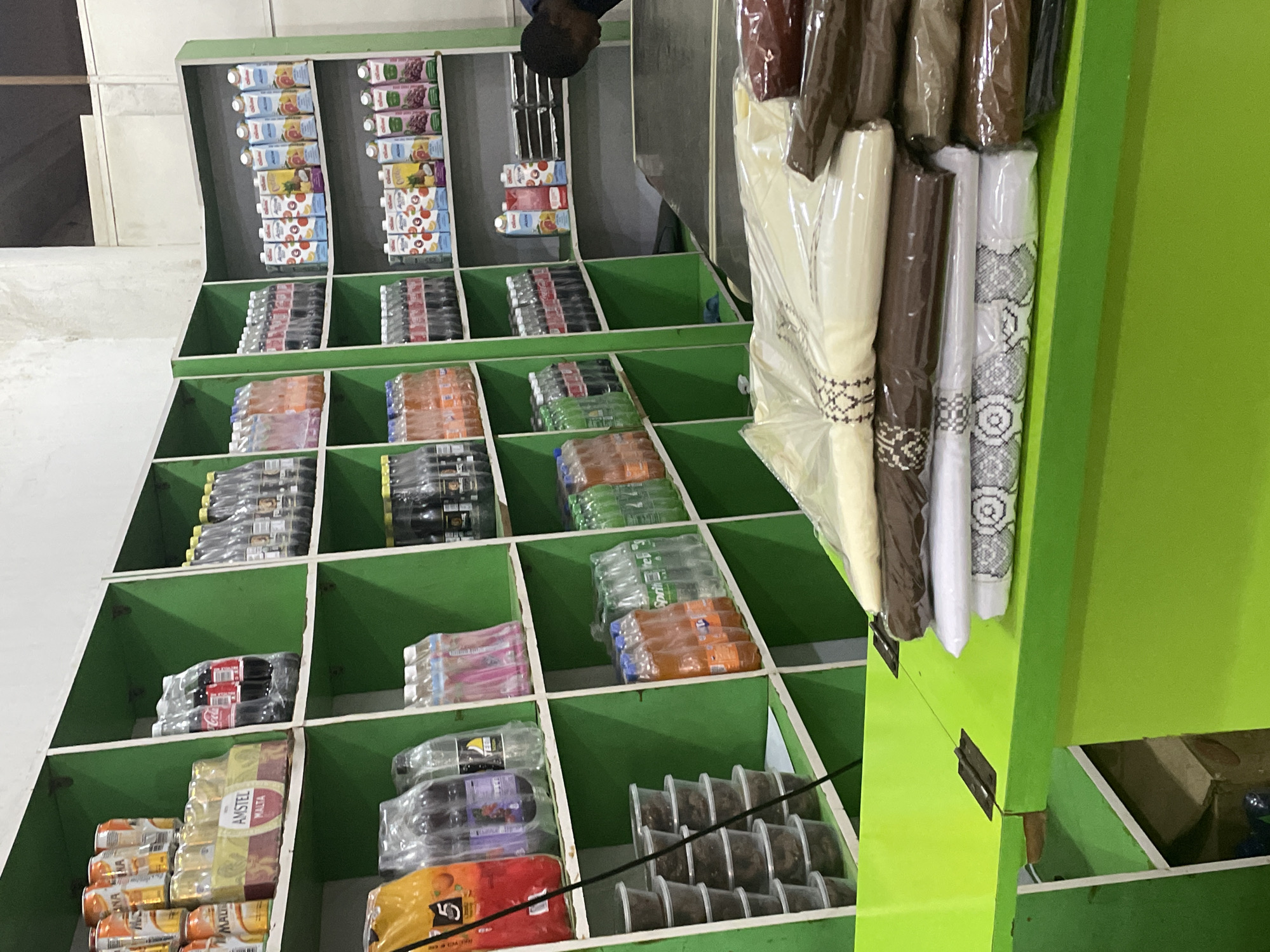 This is a media file showing the interior of a small shop, likely a convenience store or a kiosk, in Barden Yana, Nigeria. The setting is organized with green shelving units displaying various products. The items are predominantly food and beverages, including juice boxes, bottled water, soft drinks, and canned beverages. Some shelves also hold packaged food items in plastic containers.
In the upper left corner of the image, a person's shoulder and head are visible, suggesting a shopkeeper or customer is present but not fully in frame. The lighting appears to be artificial, indicating the activity is taking place indoors, possibly during the day or evening. The overall impression is of a well-stocked local store.
Some visible text includes "Amstel Malta" on a yellow and red package of canned drinks. Other text on juice boxes and bottles is too small or blurry to be clearly legible. The general atmosphere of the scene is one of commerce and everyday retail activity.