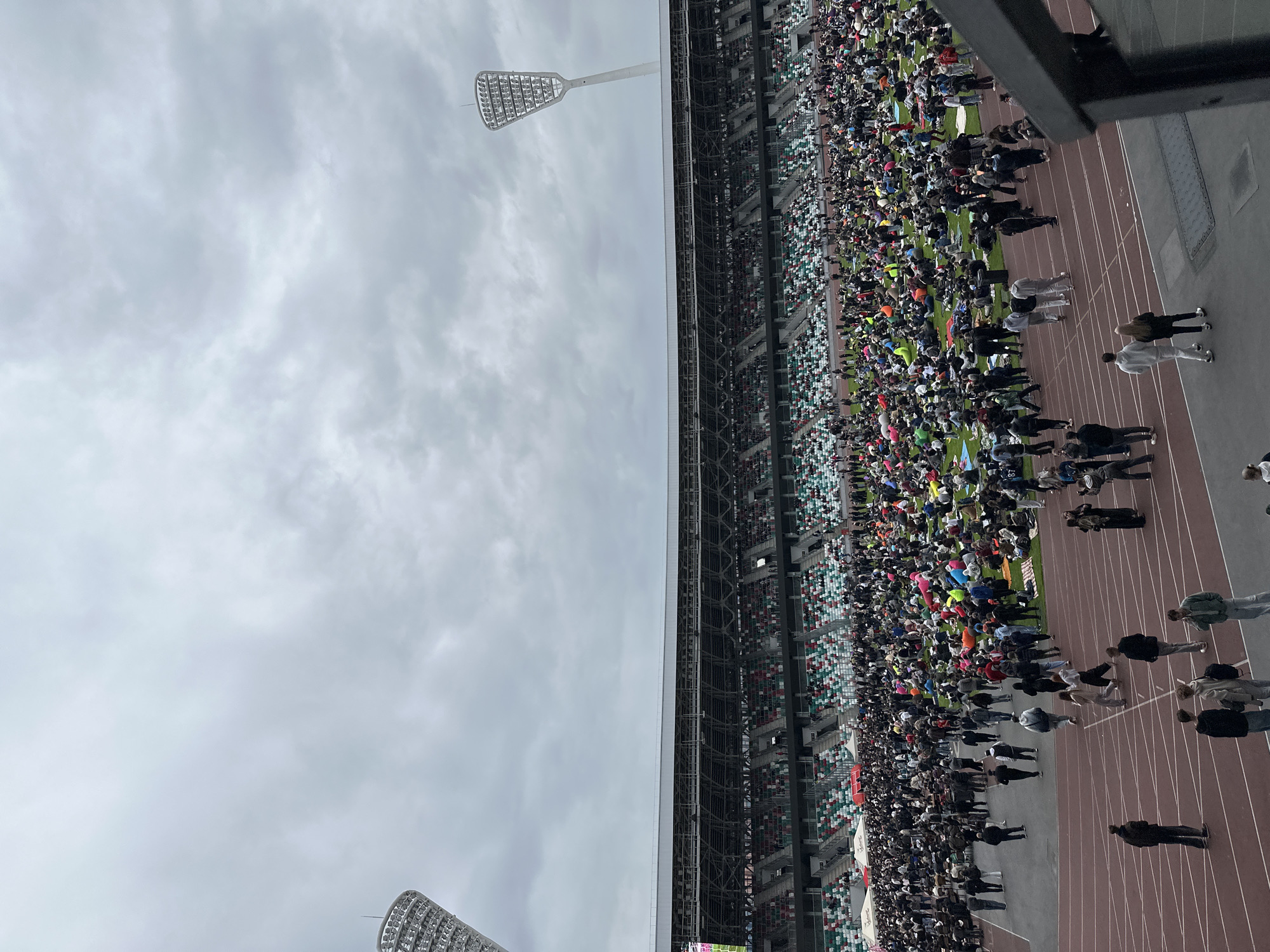 The image shows a crowded stadium in Minsk, Belarus, likely during an event. The stadium is full of spectators seated in the stands, and there is a large number of people walking around on the track surrounding the field. Stadium lights are visible overhead, and a cloudy sky is above. The people on the track appear to be predominantly adults, with some in casual clothing. There are brown lanes on the right side of the image.
