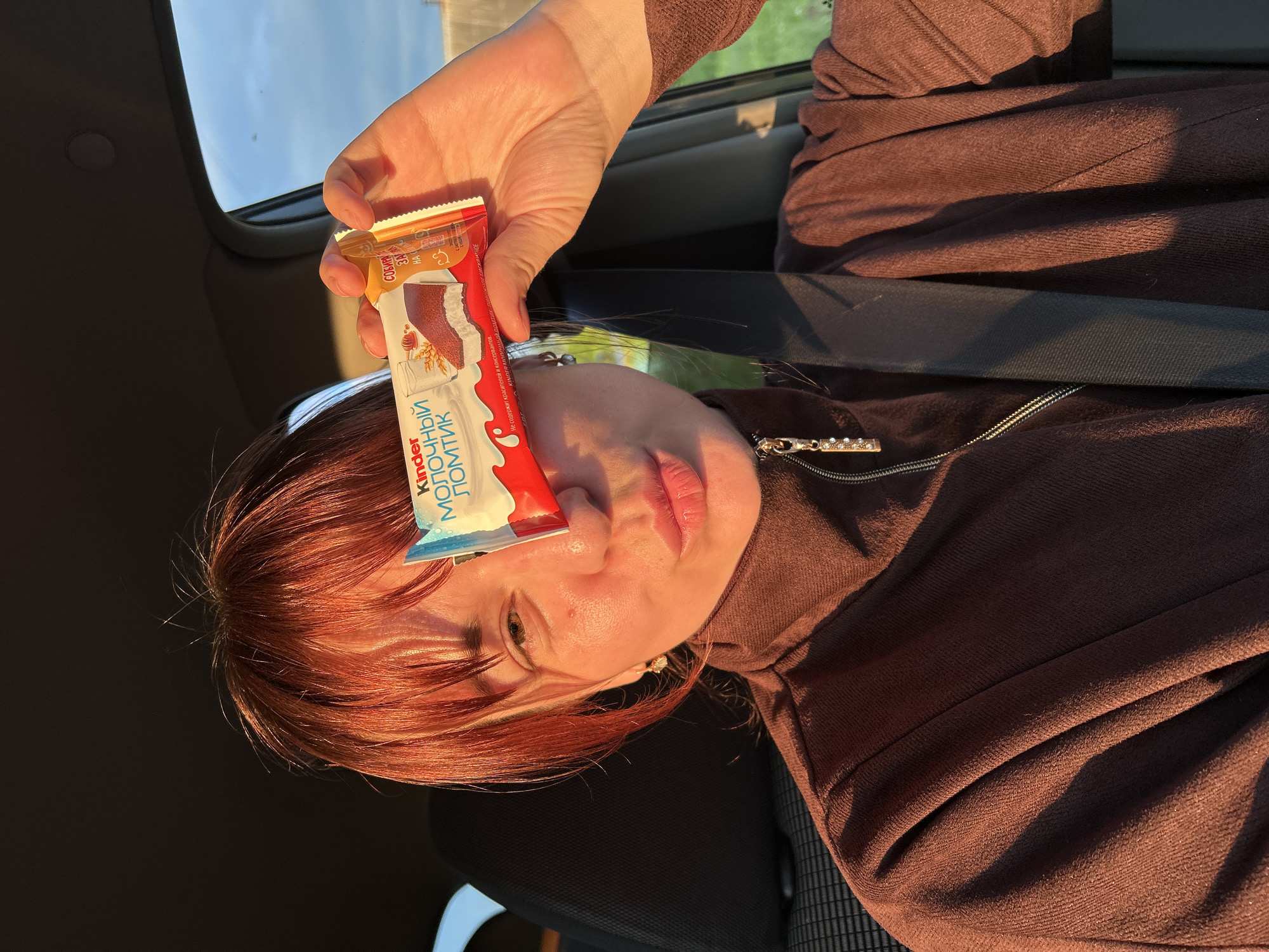 A person with reddish-brown hair is shown inside a vehicle, likely a car, holding a Kinder chocolate bar close to their face. The image is taken in portrait orientation but appears to be rotated sideways, with the person looking towards the viewer. Their face, hand, and hair are illuminated by warm, golden hour sunlight streaming through the car window, creating strong shadows and highlights. The person is wearing a brown zippered top and a seatbelt is visible across their chest. Through the car window, a clear blue sky and some distant greenery are visible, indicating an outdoor setting.
The main activity is the person presenting or posing with the Kinder chocolate bar. Their expression appears neutral, with their lips slightly parted.
Notable details include the time of day, evident from the soft, warm light characteristic of sunset or sunrise. The location is an interior of a car, suggested by the seatbelt, window frame, and seat fabric.
Visible text on the Kinder packaging includes: "Kinder", "МОЛОЧНЫЙ ЛОМТИК" (Milk Slice), "СОБИРА" (likely part of a larger word), and "Не содержит красителей и консервантов." (Does not contain dyes and preservatives.). The scene is set in Фоки, Russia.