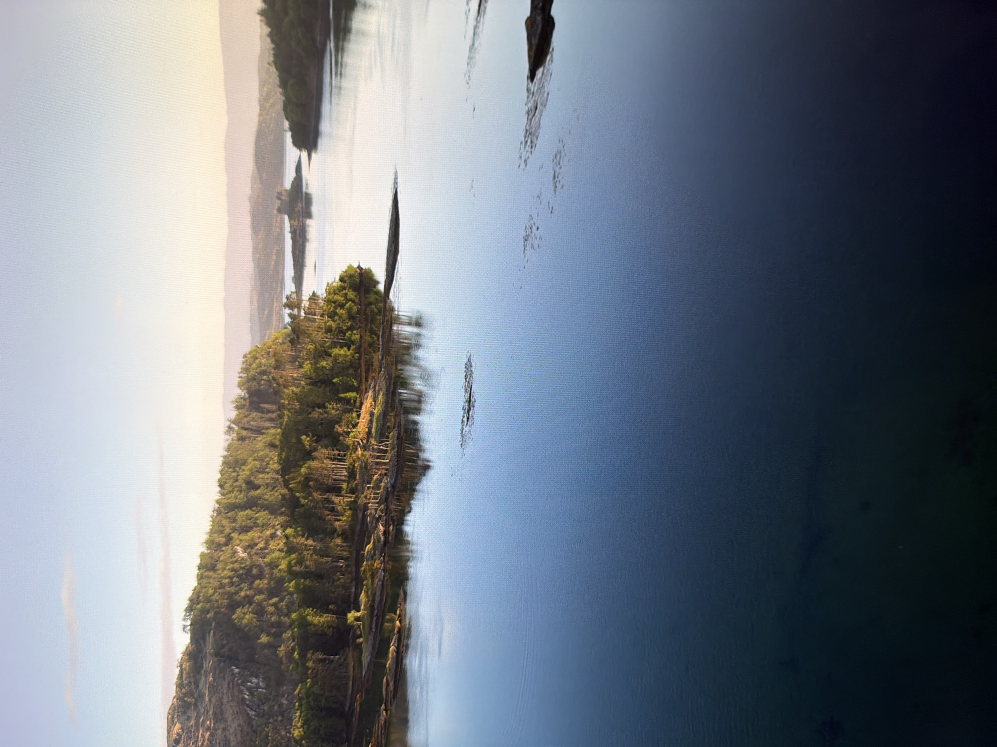 This is an aerial, wide-angle shot of a serene body of water, possibly a river or a lake, reflecting the soft light of a hazy sunrise or sunset. The foreground is dominated by the dark, tranquil water, which transitions to a lighter, more luminous blue as it stretches towards the horizon. Scattered dark shapes, possibly submerged rocks or debris, break the glassy surface in the mid-ground.
To the left, a densely forested hillside slopes down to the water's edge, with a mix of deciduous and coniferous trees. The trees are illuminated by the low-angle sun, creating a dappled effect of light and shadow. Rocky outcrops and smaller islands with sparse vegetation are visible along the shoreline.
In the distance, the water widens and curves, leading towards a small island topped with a stone structure resembling a castle or ruin. Further beyond, rolling hills fade into the hazy horizon, painted in soft pastel shades of blue and pink. The sky is a pale, gradient blue, with a faint wisp of cloud.
The scene is devoid of any human presence or activity. The overall impression is one of natural beauty and peaceful isolation.
This image does not depict any location in Kyiv, Ukraine.