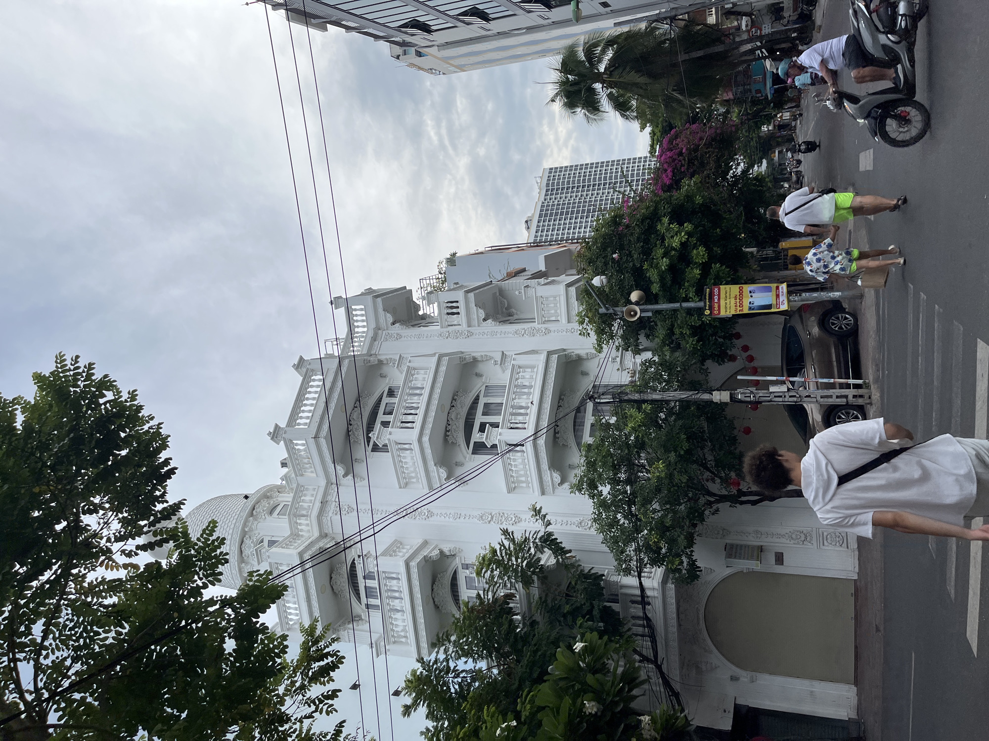 The image captures a street scene in Nha Trang, Vietnam, under a cloudy sky. In the foreground, a young person wearing a white hoodie and light-colored pants walks away from the camera, crossing a pedestrian street. To the right, a man in a white polo shirt and neon green shorts holds hands with a child wearing a patterned shirt and matching shorts, also crossing the street. Behind them, a man on a scooter wears a blue helmet and a white polo shirt. The background features a distinctive white building with ornate balconies and a domed roof, likely an older architectural style. Adjacent to it, a modern, taller building with a glass facade stands out. Further down the street, more buildings and palm trees are visible, along with several motorbikes and cars. A yellow advertisement sign for "realme" is prominently displayed on the left side of the street. The overall scene depicts a typical urban street in Nha Trang, with a blend of traditional and modern architecture and active street life.