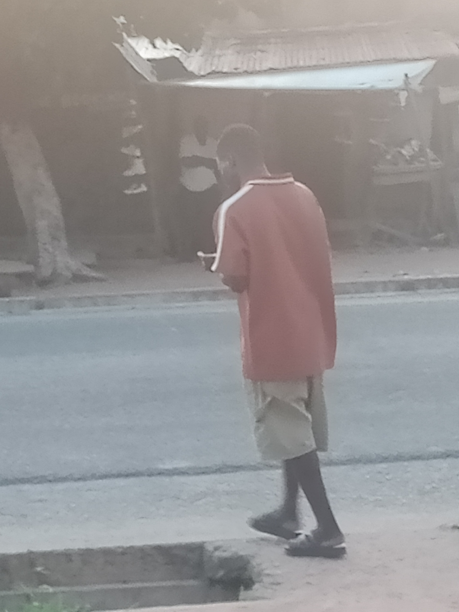 The image depicts a person walking along a street, possibly in Jalingo, Nigeria. The individual is wearing a reddish-orange polo shirt with white trim on the collar and sleeves, and light-colored shorts. They are walking with their left leg forward, and their right leg slightly behind. The person appears to be holding a light-colored object in their right hand.
The environment suggests an outdoor urban setting. A paved road is visible in the foreground, with a concrete curb separating it from a dirt or sandy area. In the background, there are structures that resemble market stalls or shops with corrugated metal roofs. A tree with a rough trunk is to the left of the frame. The overall lighting suggests it is daytime, but the image quality is low, making finer details blurry. The scene is captured from a slightly elevated perspective, looking down at the person.