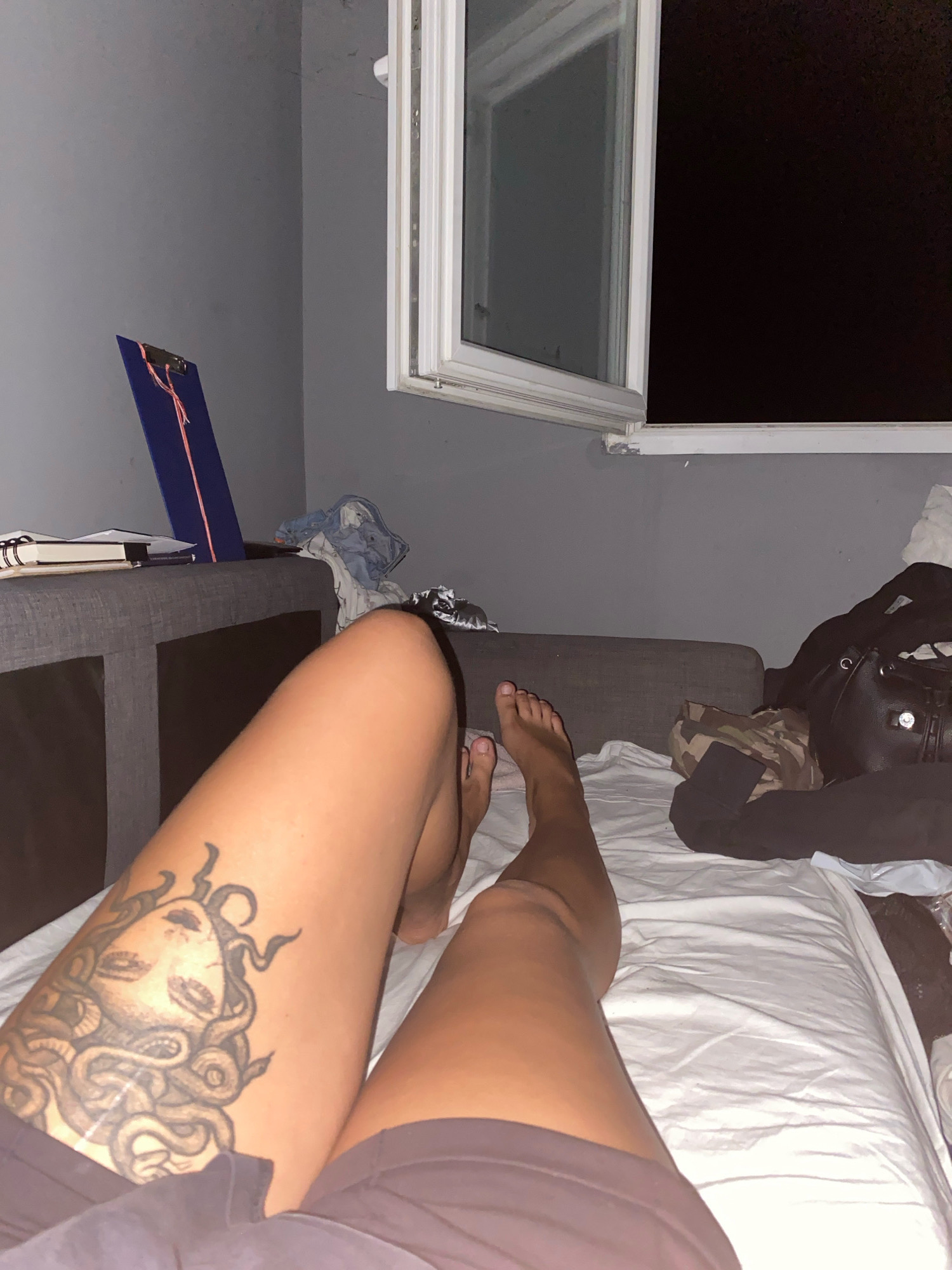 The image depicts an indoor scene, likely a bedroom, as suggested by the presence of a bed with white sheets. The room has grey walls. A window with a white frame is visible, and through it, a dark exterior is seen, indicating it is nighttime.
In the foreground, a person's legs are prominent. The left leg has a tattoo of Medusa on the thigh. The person is wearing dark shorts. To the right, the person's feet are visible.
On a grey couch to the left of the image, a blue clipboard with pink string is resting. Some folded clothing is also present on the couch. To the right of the couch, near the bed, there are various items including a black backpack and some camouflage-patterned fabric.
The overall impression is a casual, lived-in space. There is no specific indication of the city of Kučiće, Croatia, within the visual content of the image.