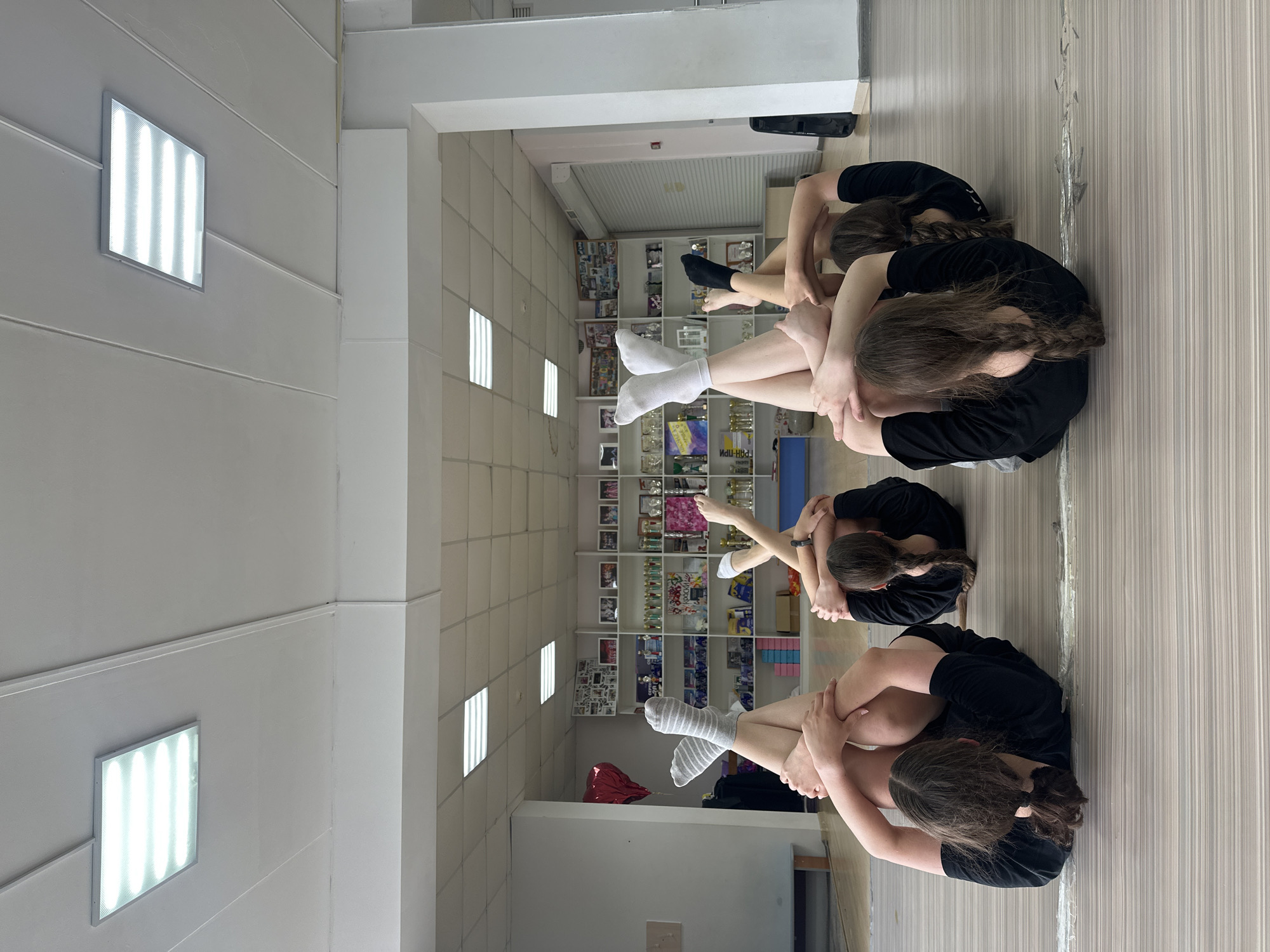 Four young girls are lying on the floor in a dance studio, each in a similar yoga-like pose. Their legs are bent and crossed, with their arms wrapped around their knees. They are all wearing black tops and athletic bottoms. The studio has a polished wooden floor and white walls. Fluorescent lights are visible on the ceiling. In the background, shelves are filled with trophies and awards, suggesting the room is used for dance competitions or performances. The overall scene depicts a group of young athletes practicing their routine in a dedicated space. The location is a dance studio in Votkinsk, Russia.