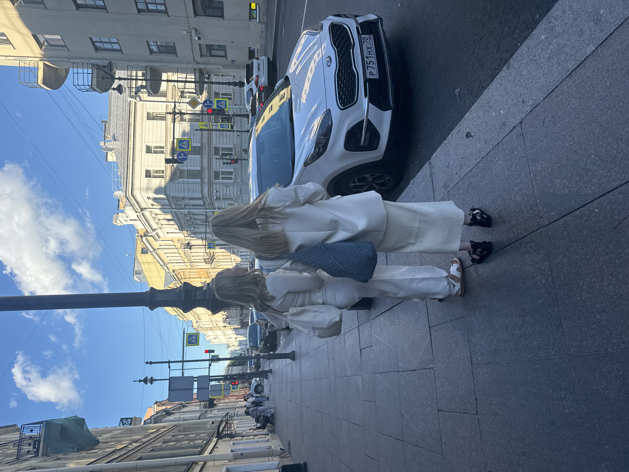 Two women wearing white outfits are standing on a sidewalk in Saint Petersburg, Russia. The woman on the left is wearing white pants and a white top, and she has a white tote bag. The woman on the right is wearing a white skirt and a white blazer, and she has a blue woven bag. They are facing away from the camera, looking towards the street. A white Kia Sportage is parked on the street in front of them. The street is lined with buildings with classical architecture. There are traffic lights and pedestrian crossing signs visible. The sky is blue with some clouds. The overall scene depicts a daytime urban environment.