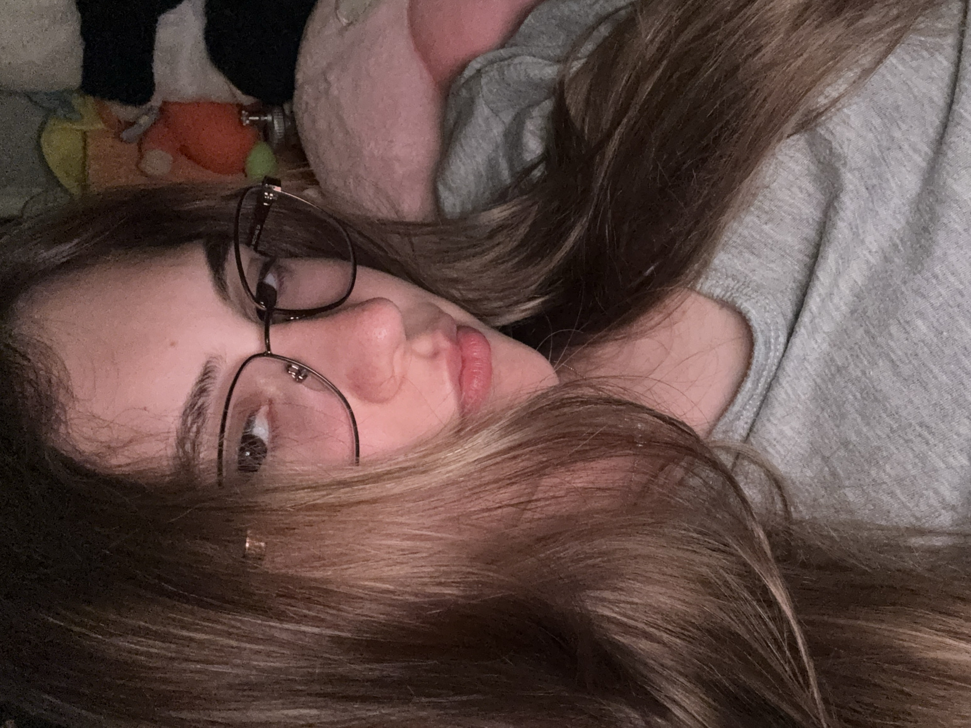 The image features a close-up, rotated portrait of a person, likely a young woman, lying down indoors. Her head is positioned on the left side of the frame, with her body extending to the right. She has long, wavy brown hair that spreads across much of the image. She is wearing round, thin-framed glasses, and her right eye is visible, looking towards the viewer. Her lips are slightly parted, revealing a neutral expression.
She is wearing a light grey top or sweater with a visible textured or knitted pattern. In the background, behind her head, several soft objects or plush toys are discernible in muted colors, including pink, orange, black and white, and hints of green and yellow, suggesting a comfortable, possibly bedroom or living room setting. The lighting is dim, indicating it might be evening or nighttime. No text is visible in the image. The overall impression is one of relaxation or contemplation in a casual indoor environment.
