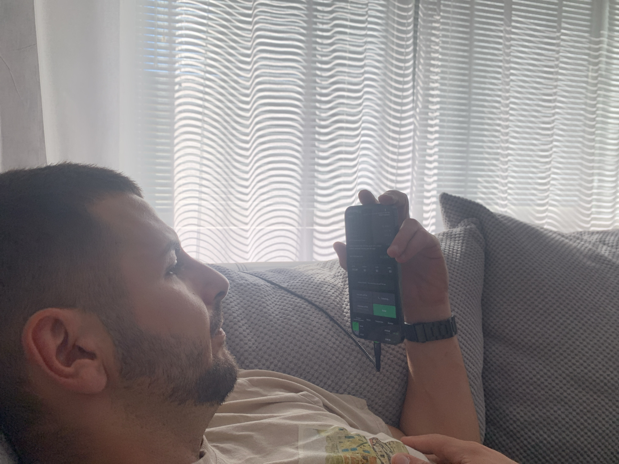 A man is lying on a gray, textured sofa in front of a window covered with wavy blinds. He is holding a smartphone in his right hand and appears to be looking at its screen. The phone screen displays a trading app with figures in green. The man has short, dark hair and a beard. He is wearing a tan-colored t-shirt and a dark-colored watch on his wrist. The background is brightly lit. There is no clear indication of the specific location in Lovech, Bulgaria, as the image does not contain any identifiable landmarks or indicators of place.