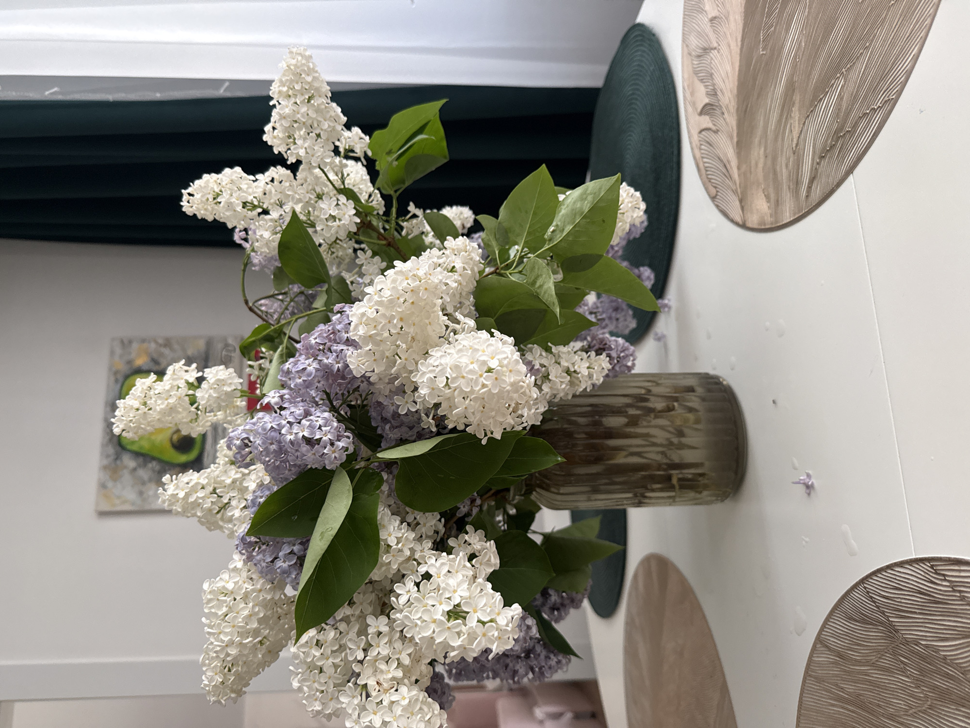 A vase filled with a bouquet of white and lilac-colored lilacs rests on a light-colored surface. The vase appears to be fluted with a horizontal pattern. Droplets of water are visible on the surface around the vase. In the background, there is a glimpse of a painting on the wall with green and grey hues. Behind the flowers is a dark green fabric. The surface the vase is sitting on has decorative concentric circles. The location cannot be determined from the image.