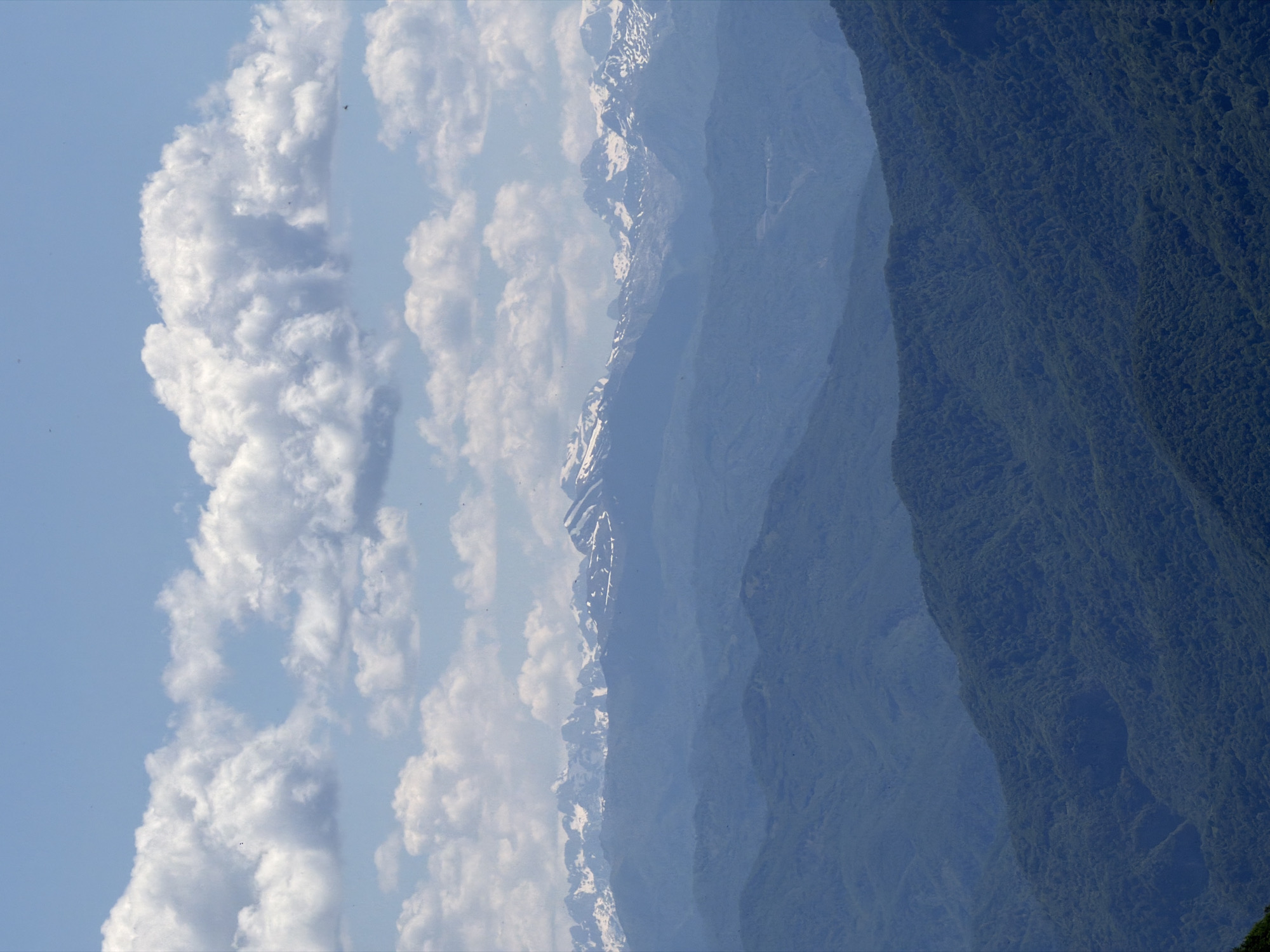 The image displays a landscape composed of layered elements: a sky filled with fluffy white clouds, snow-capped mountain peaks, and a dense, dark green forest. The perspective is from a high vantage point, looking down at the scene. The clouds are illuminated, contrasting against the blue sky. The mountains fade into a hazy blue, with visible patches of snow clinging to the higher elevations. The forest on the right appears dense and textured. Location: Unknown, Montenegro.