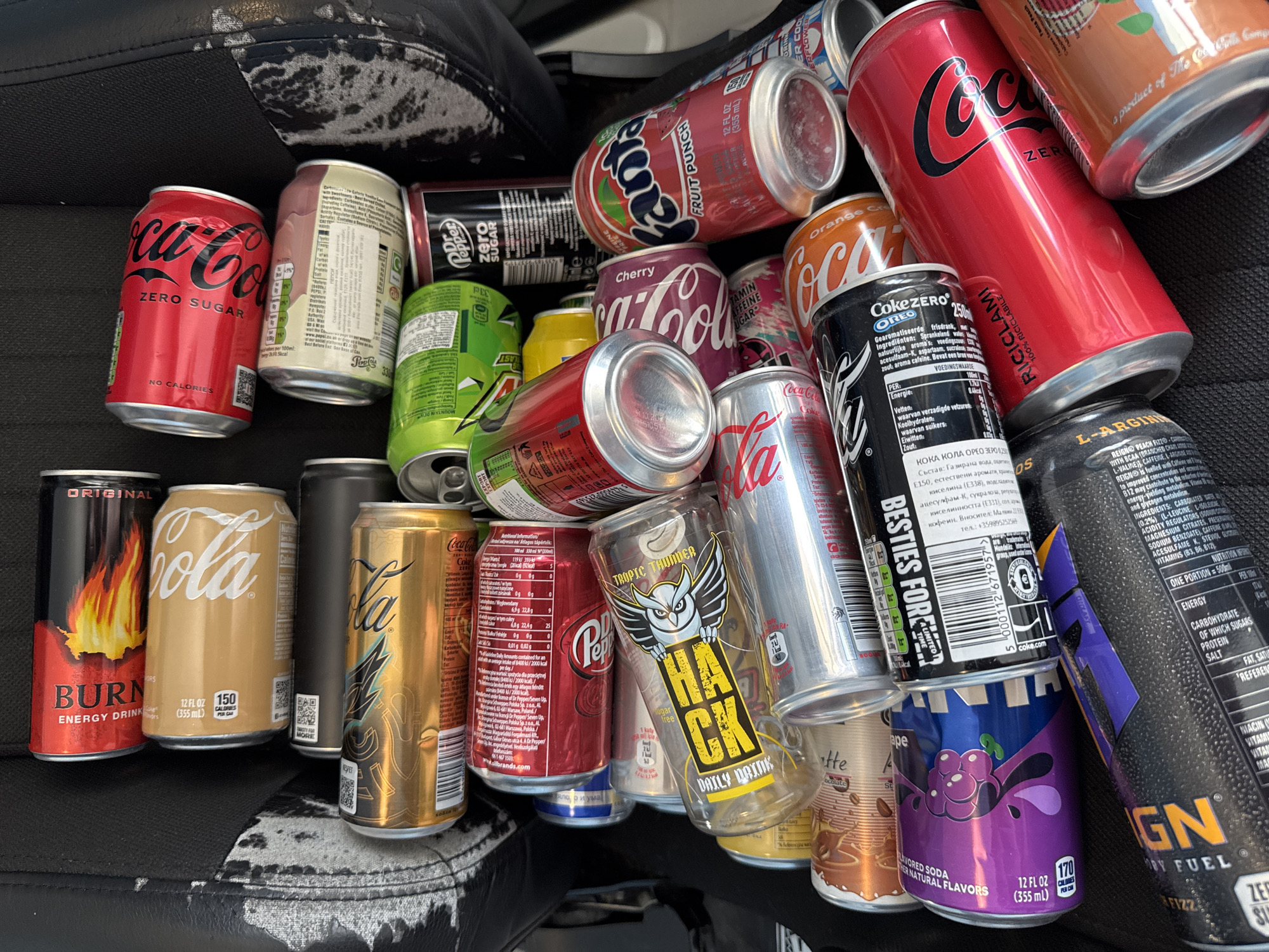 The image shows a collection of approximately 25 empty soda and energy drink cans piled on a black surface, likely a car seat given the visible distressed leather pattern. The brands are diverse, including Coca-Cola (various flavors, including Zero Sugar, Cherry, and Orange Vanilla), Fanta (Fruit Punch), Dr. Pepper Zero, Burn Energy Drink, Hack Daily Dose (Tropic Thunder), Grape soda, Coke Zero Oreo, and Reign energy drink (Peach Fizz). The labels are predominantly in English, with some text in languages like Bulgarian (on the Coke Zero Oreo can, noting the importer as Mali 2283 Sofia) and other European languages (nutrition information). The cans appear to be discarded or collected for recycling. The location cannot be determined from the image.