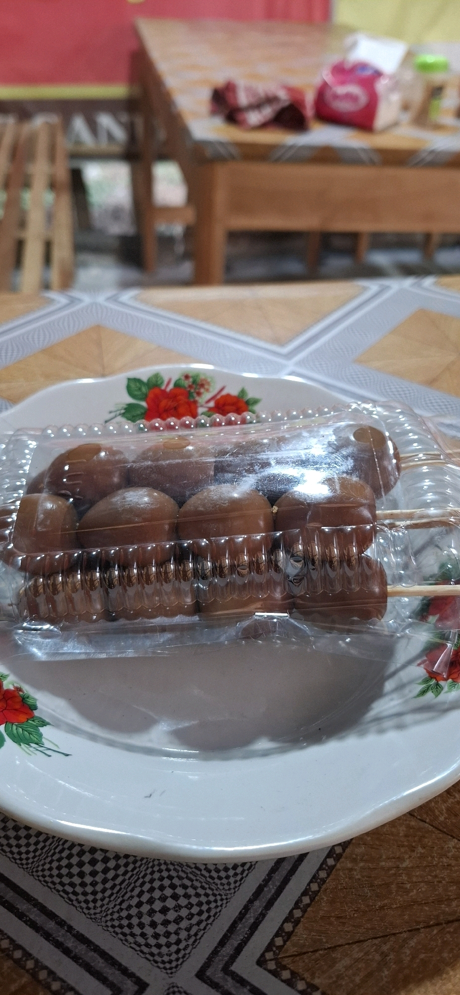 The image shows a close-up of a transparent plastic container filled with several chocolate-covered candies on skewers, placed on a white plate with red floral decorations. The plate is situated on a patterned tablecloth. In the background, a wooden table with a red cloth and a tissue box is visible. The scene appears to be indoors, possibly in a home or small eatery. The specific location in Colomadu, Indonesia is not determinable from the visual information alone.
