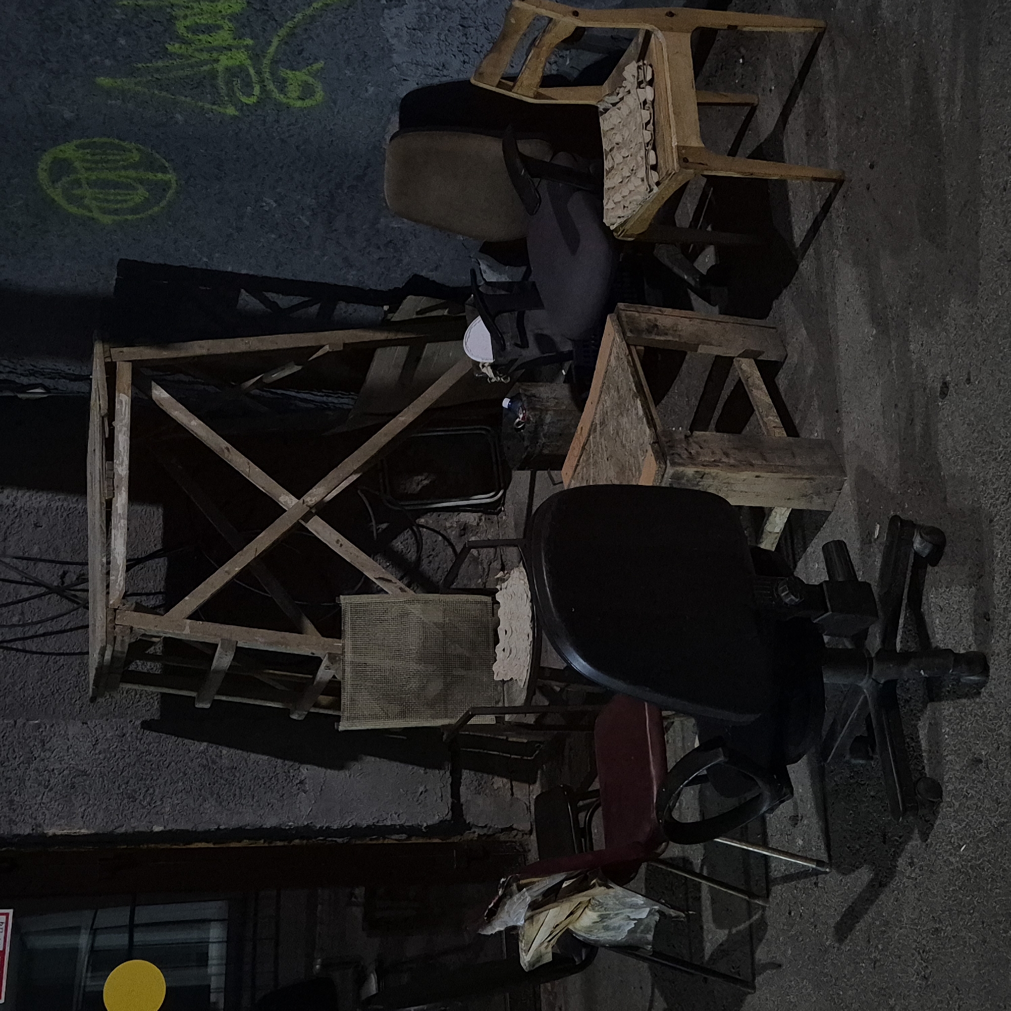 The image depicts an outdoor scene with several chairs, a wooden structure resembling a makeshift scaffold, and some scattered debris. The setting appears to be an alleyway or a neglected outdoor space, possibly in the city of Kemerovo, Russia, given the context provided. The lighting suggests it is nighttime or a very dimly lit area.
In the foreground, a black office chair with wheels is prominently visible. To its left, a red chair is partially obscured. Behind these chairs, a small wooden table is placed. Further back, a brown office chair sits near a wooden chair with an egg carton on its seat. A tall, wooden structure with crisscrossing supports stands in the background. The wall behind the structure is textured and has some yellow graffiti. The ground is paved with a rough material. There are no people visible in the image. The overall impression is one of disuse and clutter.