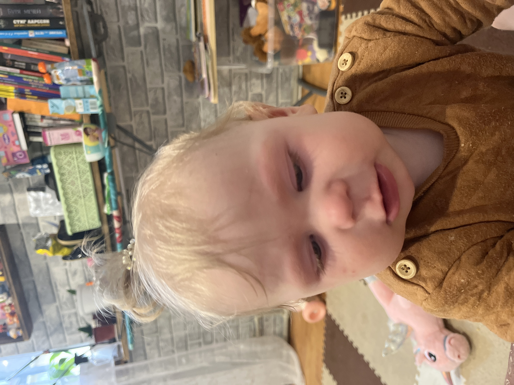 This portrait-style image, taken from a slightly upward angle, captures a fair-skinned baby or young child with light blonde hair, looking directly towards the viewer. The child wears a textured, brown long-sleeved top, featuring two light wooden buttons on the right shoulder. A small, elegant white hair clip with pearl-like embellishments is visible in the child's hair, positioned towards the back of their head. The child has a playful expression, with a subtle smirk and their tongue just peeking out between their lips.
The setting is indoors, likely a home environment such as a child's room or play area. In the background, to the child's left, a bookshelf is filled with books and various small items. Several book spines display Cyrillic script, with fragments like "СТУРМ" (Storm) and hints of "БАРСЕЛОНА" (Barcelona) or "СССР" identifiable, strongly suggesting a Russian context, which aligns with the provided location of Vesyoloye, Russia. Below the books, various packaged items, possibly snacks or drinks, are stacked. The wall behind the bookshelf features a grey brick or stone pattern.
The floor beneath the child is covered with interlocking foam play mats in alternating shades of brown and beige. A pink stuffed animal, resembling a unicorn or a similar creature, lies on the floor to the child's right. The lighting indicates an indoor daytime scene, and the child's demeanor appears content and engaged.