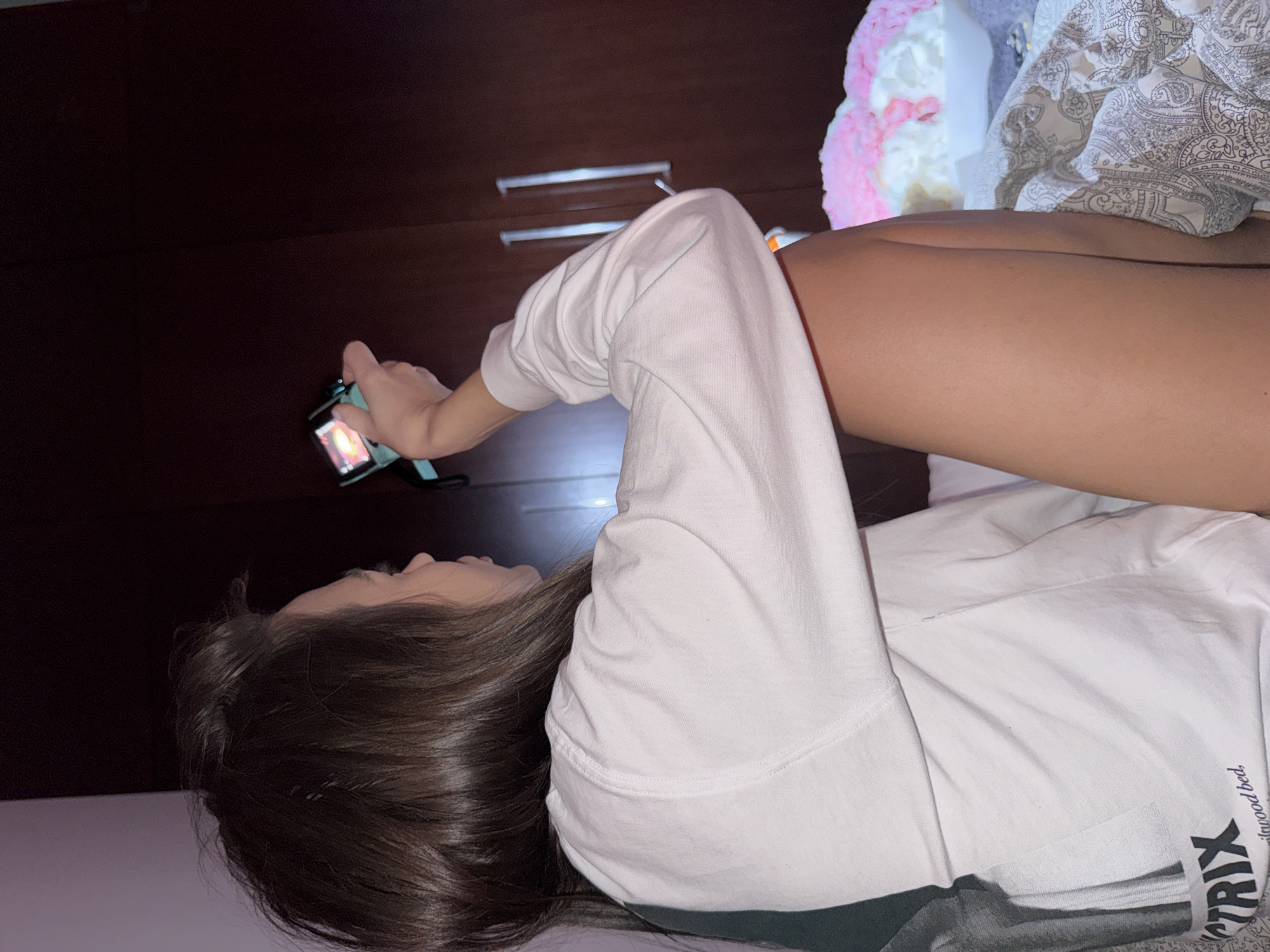 This indoor image, rotated 90 degrees counter-clockwise, depicts a young person, likely female, with long, dark brown hair, lying on their back in what appears to be a bedroom in Warsaw, Poland. The room is dimly lit, suggesting evening or nighttime.
The subject is wearing a white long-sleeved shirt or sweatshirt, with a visible darker lower section, and her bare legs are exposed. She is focused on a smartphone, held in her left hand, which is bent at the elbow. The phone's screen is illuminated, displaying bright, colorful content, and it is encased in a light blue or teal cover.
Behind her, to the left of the frame, is a dark brown wooden wardrobe or dresser featuring two silver horizontal handles. To her right and upper right, there are soft furnishings, including a textured pink and white fluffy object, possibly a pillow or blanket, and another patterned fabric, white with a grey paisley or floral design.
The person appears relaxed and absorbed in her phone activity, possibly browsing or watching content. The white shirt has visible text near the bottom right, which reads "TRIX" in large letters, and below it, partially obscured smaller text like "wood bed". The overall atmosphere is one of quiet, personal leisure.