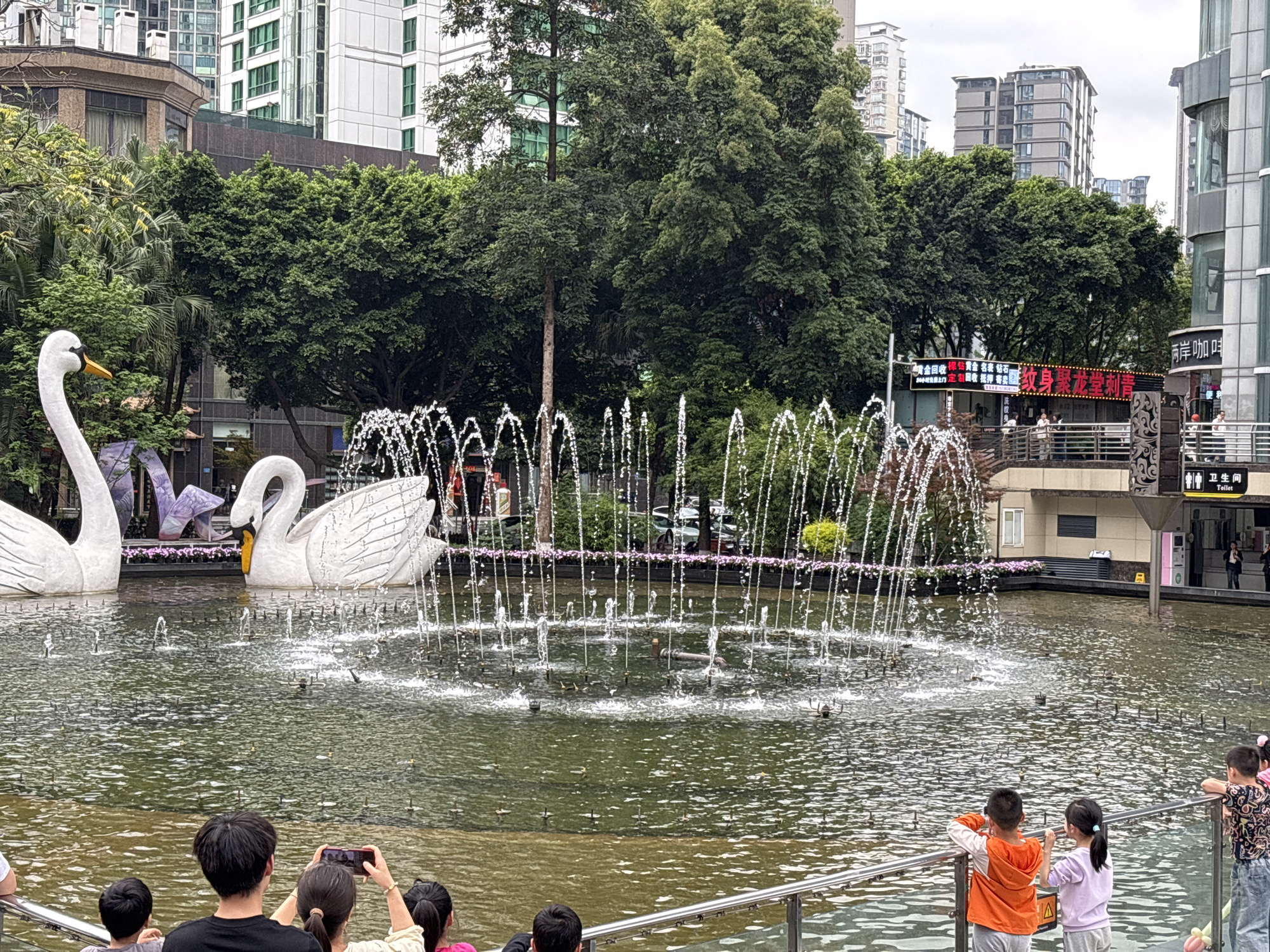 The image shows a water fountain with two large swan sculptures in a pond, surrounded by trees and buildings in the city of Liangjiang, China. The fountain is active, with multiple jets of water spraying upwards. A group of people, including several children, are observing the fountain from behind a metal railing. A woman is taking a photo of the fountain with her mobile phone. A sign in the background includes information about a tattoo parlor named "纹身聚龙堂刺青" and toilet directions.