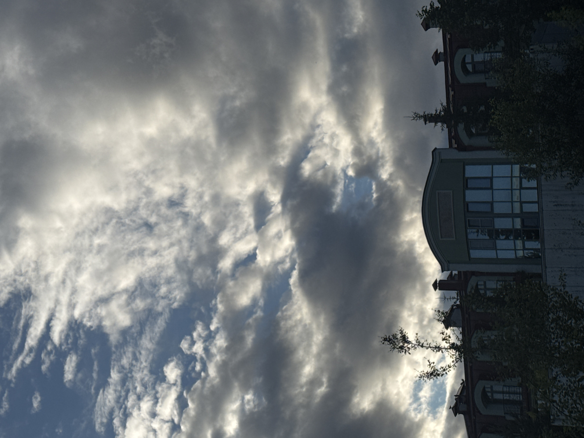 The image depicts a cloudy sky above a building in Saint Petersburg, Russia. The sky is filled with a mix of dark grey and white clouds, with patches of blue sky visible through them. The building appears to be several stories high and has a red roof. There are windows on the upper floors, some with arched tops. Trees and foliage are visible in front of the building, obscuring part of its lower sections. The overall impression is of a somewhat overcast day in an urban setting.