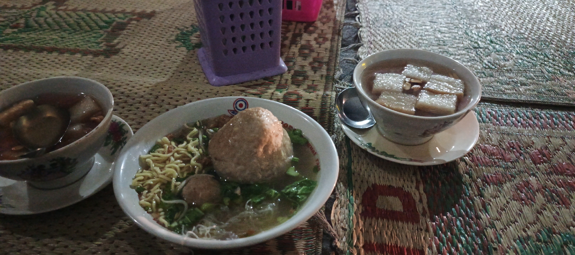 Two bowls of food are placed on a woven mat. The bowl on the left contains noodles, a large meatball, and some greens. The bowl on the right contains a broth with rectangular pieces of food. A purple plastic container is visible in the background. The setting appears to be an outdoor eatery in Godean, Indonesia, likely a bakso stall, given the presence of meatballs and noodles. The mat is likely a traditional Indonesian lesehan mat, commonly used for dining on the floor. The lighting suggests it is evening or nighttime.