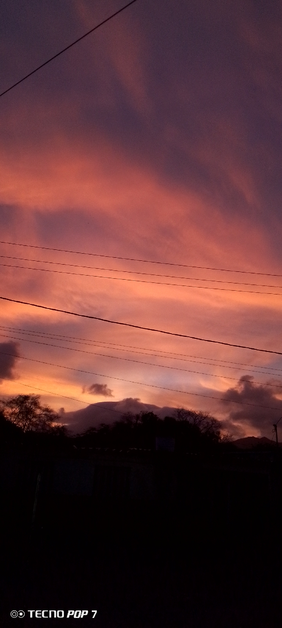 The image shows a sunset in Altagracia, Venezuela. The sky is filled with clouds in shades of orange, pink, and purple. Silhouetted mountains and trees are visible on the horizon. Power lines stretch across the sky. The foreground is dark, obscuring details of the landscape. A watermark indicating the image was taken with a TECNO POP 7 phone is present in the bottom left corner.
