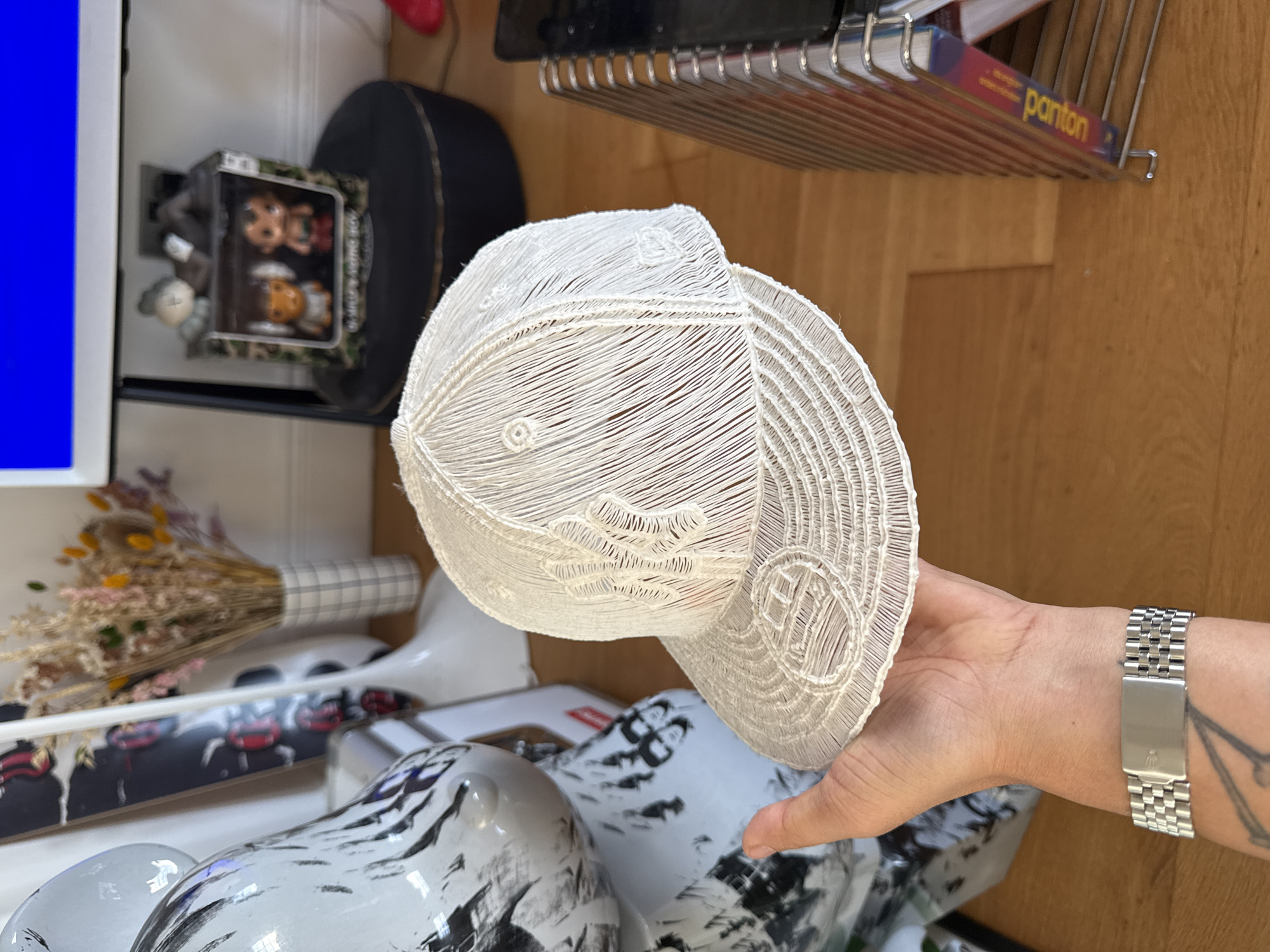 This indoor photograph, rotated 90 degrees counter-clockwise, captures a hand presenting a unique white cap against a backdrop of various decorative objects within a modern interior. The setting, possibly a home or studio, is located in London, United Kingdom.
The central subject is an intricately designed white cap, resembling a baseball or trucker style, crafted from finely woven or stitched white thread, forming a delicate lattice or mesh structure. Complex patterns, including what appears to be a stylized "K" or similar emblem, adorn its front and brim. A human hand, wearing a silver-toned metal watch with a Jubilee bracelet and a rectangular clasp, holds the cap. A portion of the forearm, featuring a black fine-line tattoo, is also visible.
The background reveals a curated collection of items. On the left, a bright blue screen is partially seen, beside which a clear display box houses collectible figures. Text on the box clearly reads "KAWS SHAQ & BIGGIE" and the number "80", with a light-colored KAWS Companion figure visible. Further right, a vase holds dried flowers and grasses. In the foreground and mid-ground, multiple irregular, glossy objects with black and white abstract patterns, reminiscent of marbling or camouflage, are present. A long, flat object with red and black graphics, resembling a skateboard, lies nearby. The floor is made of wood. Above the cap, a metal shelf or rack displays books, one of which has "panton" distinctly visible on its spine, suggesting a Pantone color guide.
The image portrays a moment of showcasing the distinctive white cap. The scene is brightly lit, indicative of daytime.