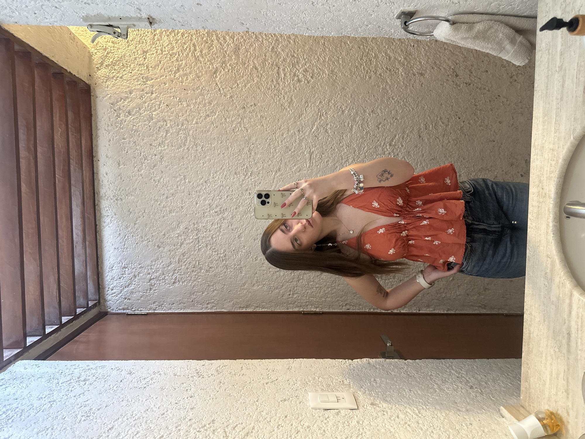 A young woman stands in a bathroom in Naucalpan de Juárez, Mexico, taking a mirror selfie with her smartphone. She wears an orange floral top and denim jeans. The bathroom has a textured white wall and a brown door. A towel hangs near the mirror. A light switch is visible below the mirror. The bathroom also has wooden blinds on one side.