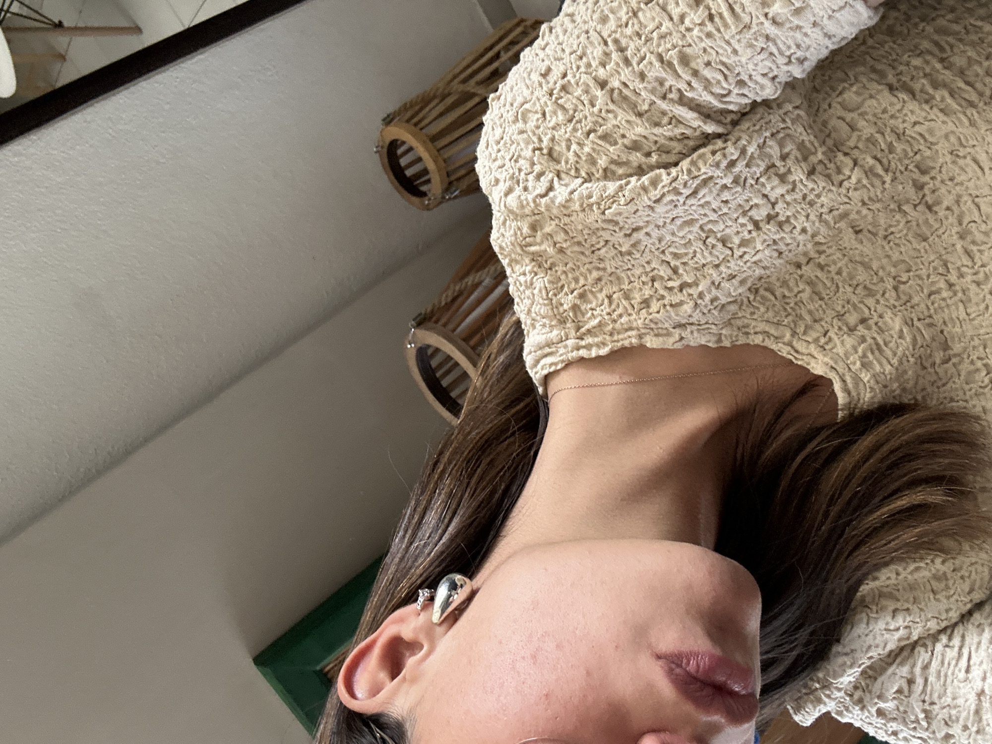The image depicts a close-up, inverted perspective of a person's face and upper torso. The individual has fair skin, dark hair, and is wearing what appears to be a textured, beige-colored garment. The person has an earring in their left ear. In the background, a white ceiling is visible, along with two cylindrical, wooden-slatted objects which are likely light fixtures. No location details are provided.