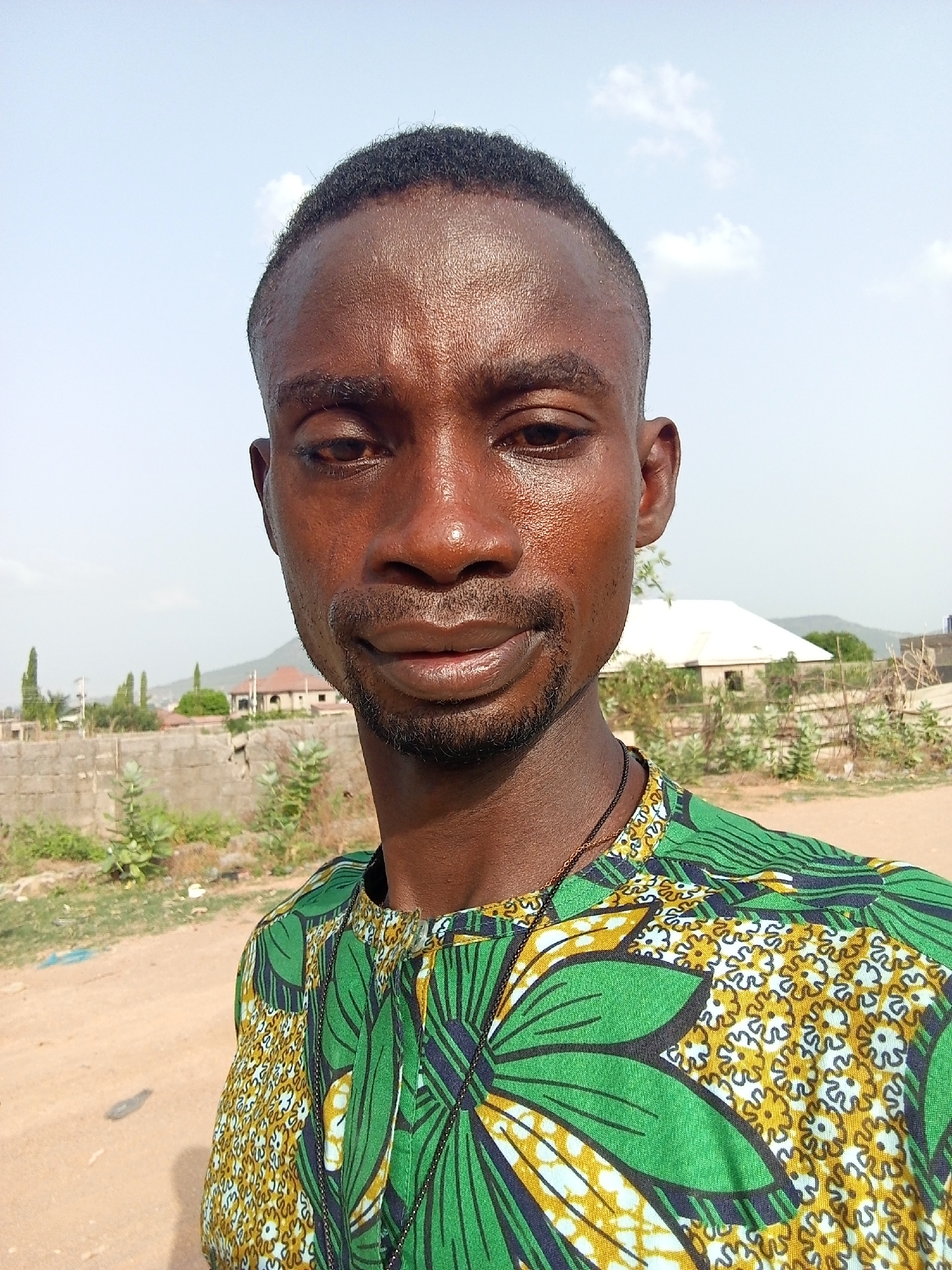 A medium shot of a man is captured under clear, bright daylight in what appears to be Otokiti, Nigeria. The man has short, dark hair and a neatly trimmed beard. His skin appears to be damp, possibly from sweat. He wears a green and yellow patterned shirt. The background shows a sparse landscape with some buildings, low walls, and vegetation. The sky is blue with a few clouds. A hill is visible in the background.
