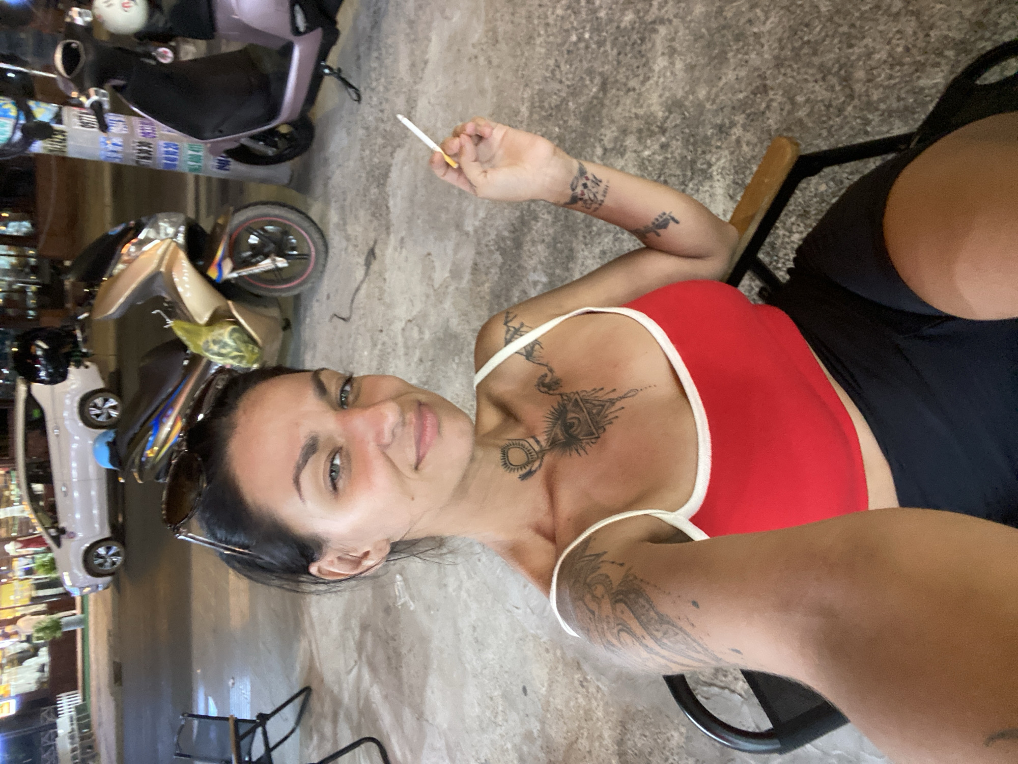 This is a self-portrait of a woman with dark hair, wearing a red top and black shorts. She is sitting in an outdoor setting, possibly at a cafe or restaurant. The background shows a street scene with motorbikes and a white car. The woman is holding a cigarette and appears to be looking at the camera with a relaxed expression. She has several tattoos visible on her arms and chest. The lighting suggests it might be evening or nighttime. The overall mood is casual and informal. The location is identified as Phu Thuy Ward, Vietnam.