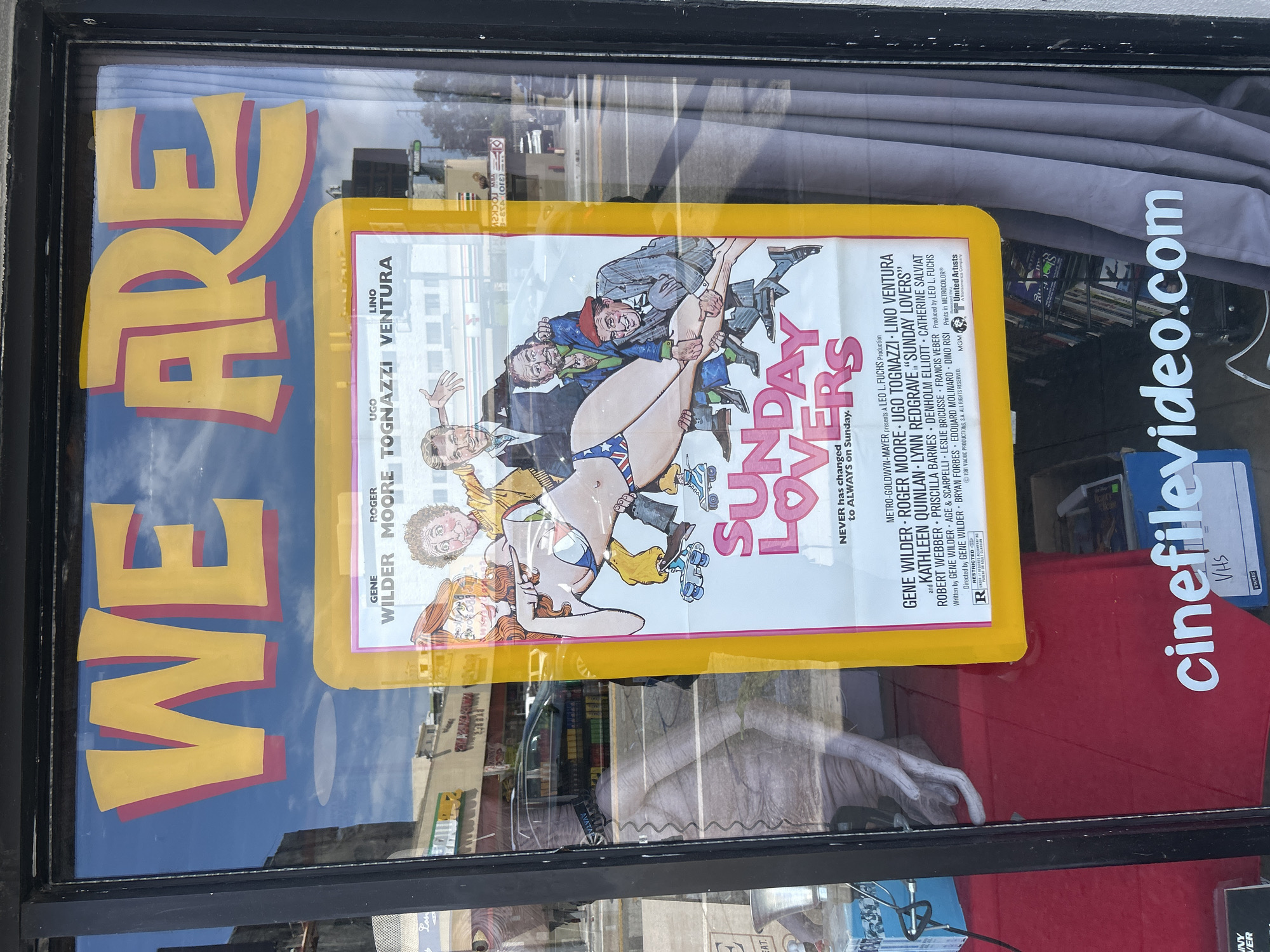The image shows a movie poster of "Sunday Lovers" (1981), affixed to a storefront window. The storefront also has "WE ARE" painted on the window. The text "cinefilevideo.com" is printed in the storefront window as well. There is no evidence in the image that can confirm the precise location in Los Angeles, CA. However, the presence of "cinefilevideo.com" is a key indicator.