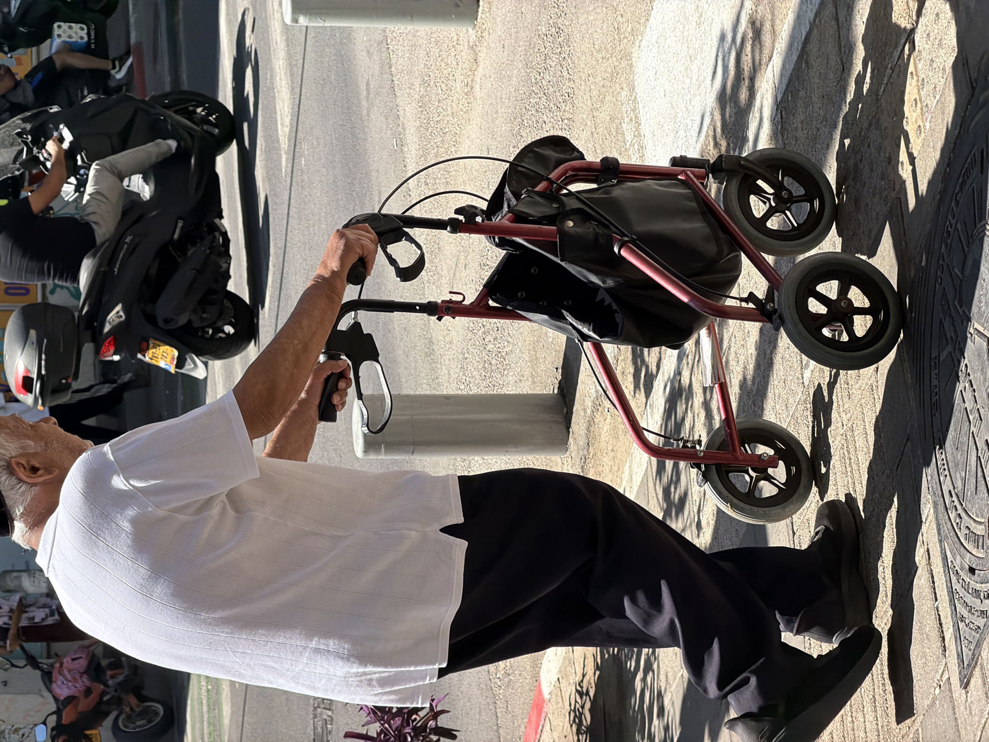 An elderly man is walking with a red folding walker on a sidewalk in Tel Aviv, Israel. He is wearing a white short-sleeved shirt, black pants, and black shoes. The walker has a black bag attached to the front. The sidewalk is made of light-colored stone and there is a manhole cover with Hebrew writing on it. A motorcycle is parked nearby and other people are visible in the background. The sun is casting shadows on the sidewalk.
