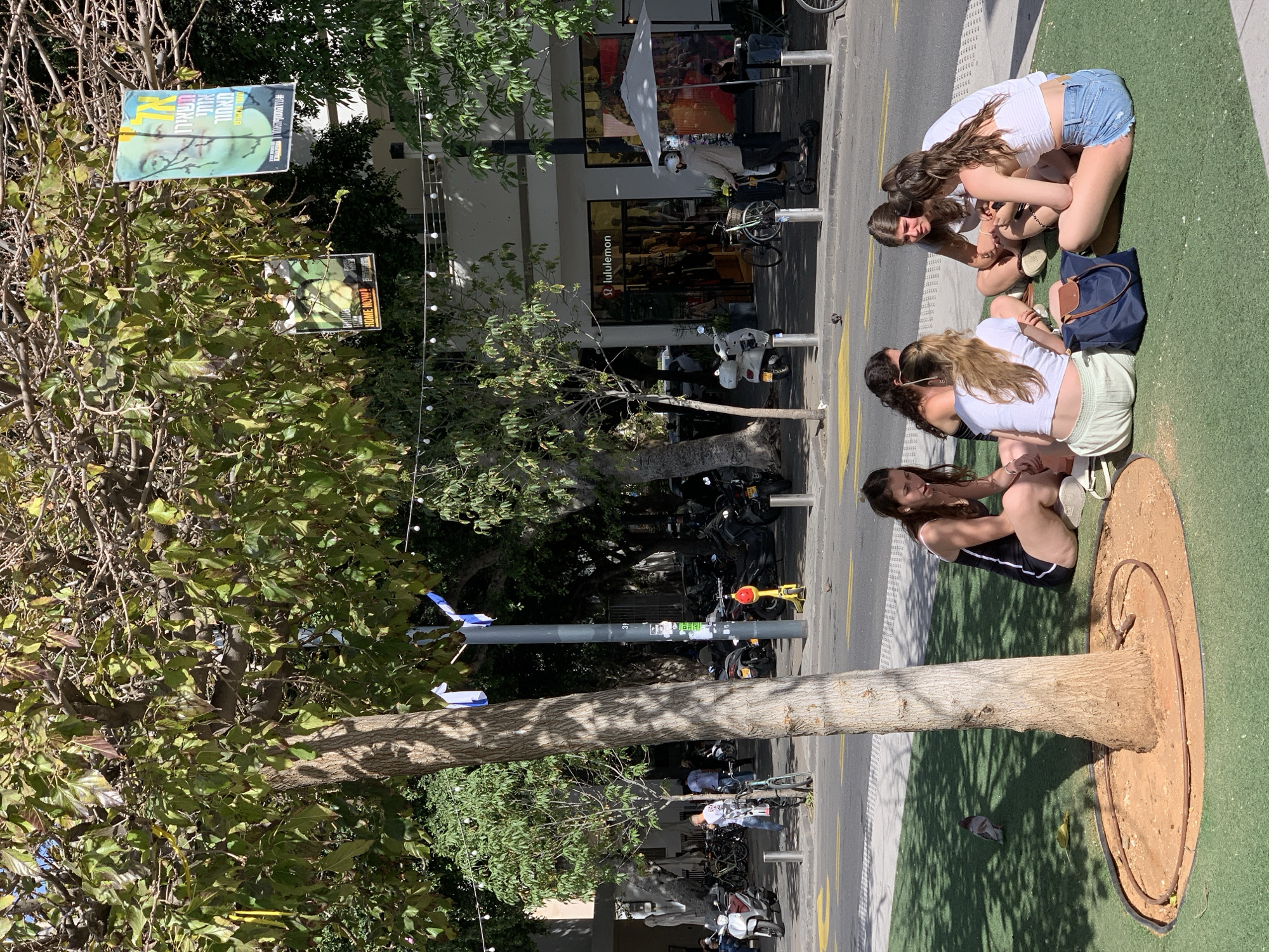 Four young women are seated on the ground near a tree in what appears to be a street in Tel Aviv, Israel. They are congregated closely, possibly engaged in conversation or using their phones. The street has pedestrian and bike lanes, and is lined with trees and buildings, including a store named "Lululemon." Bicycles and scooters are parked along the side of the street. Posters are hung on the trees. A street light pole with white decorations is next to the group.