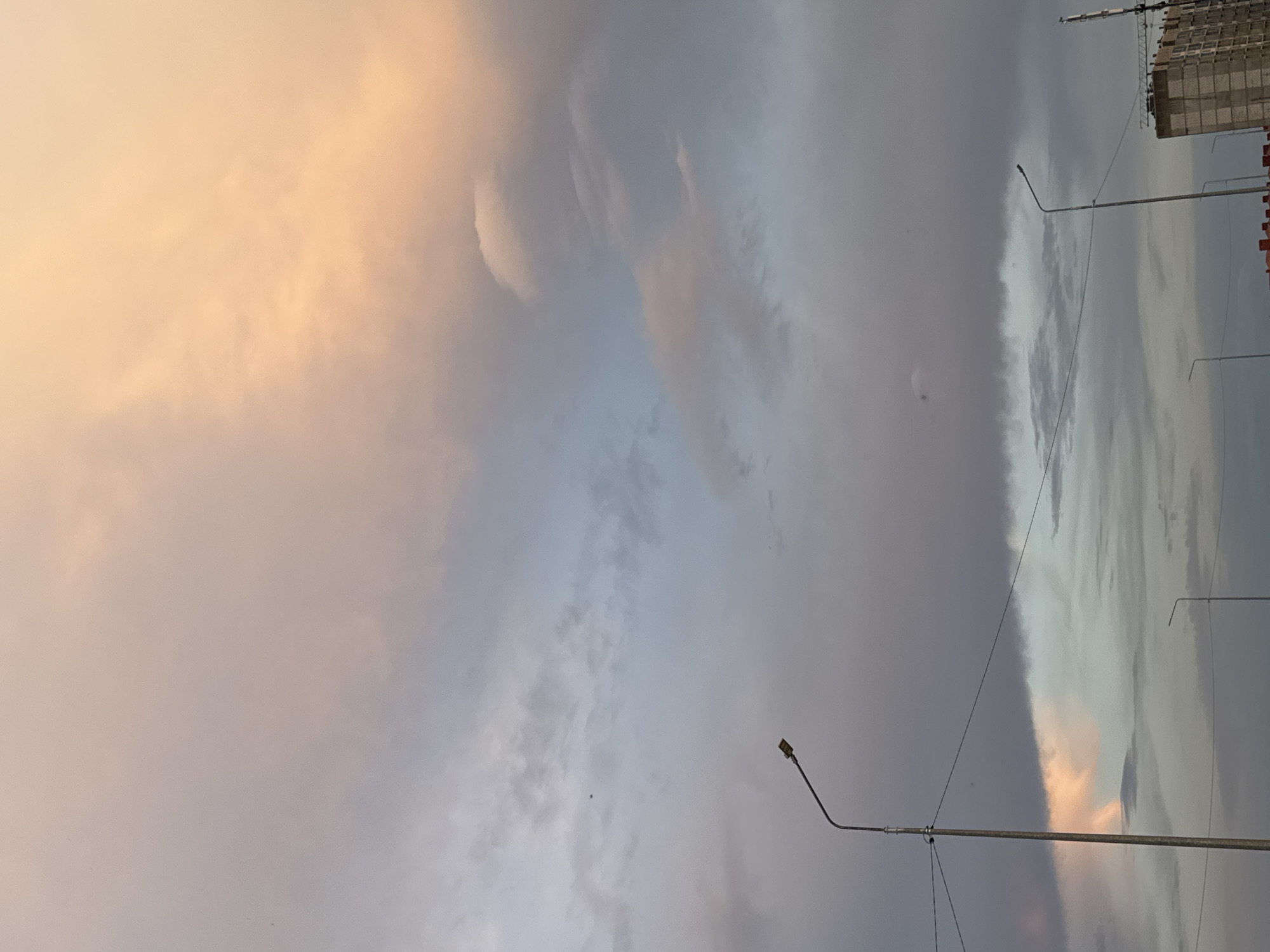 The image displays a cloudy sky with soft, diffused light, suggesting either sunrise or sunset. The clouds are varied in color, ranging from a warm, peachy hue on the left to cooler grays and blues in the center and right. This interplay of light and shadow creates a dramatic and somewhat moody atmosphere.
In the background, on the right side of the frame, a tall building under construction is visible, indicating an urban or semi-urban setting. The building appears to be made of concrete and brick, with scaffolding or construction materials present. Two streetlights with a modern design are also visible. One is in the foreground, a tall pole with a single lamp at the top, and another is further in the background, smaller and less distinct. The presence of these man-made structures contrasts with the natural element of the sky.
There are no people or animals clearly visible in the image. The activity is passive, focused on the sky and the overall scene. The time of day is likely early morning or late evening due to the lighting conditions and the warm tones in the clouds. The weather appears to be overcast or partly cloudy, with the potential for changing conditions given the dramatic cloud formations. No text is visible in the image. The overall impression is one of a serene yet somewhat somber urban landscape at the edge of the day.