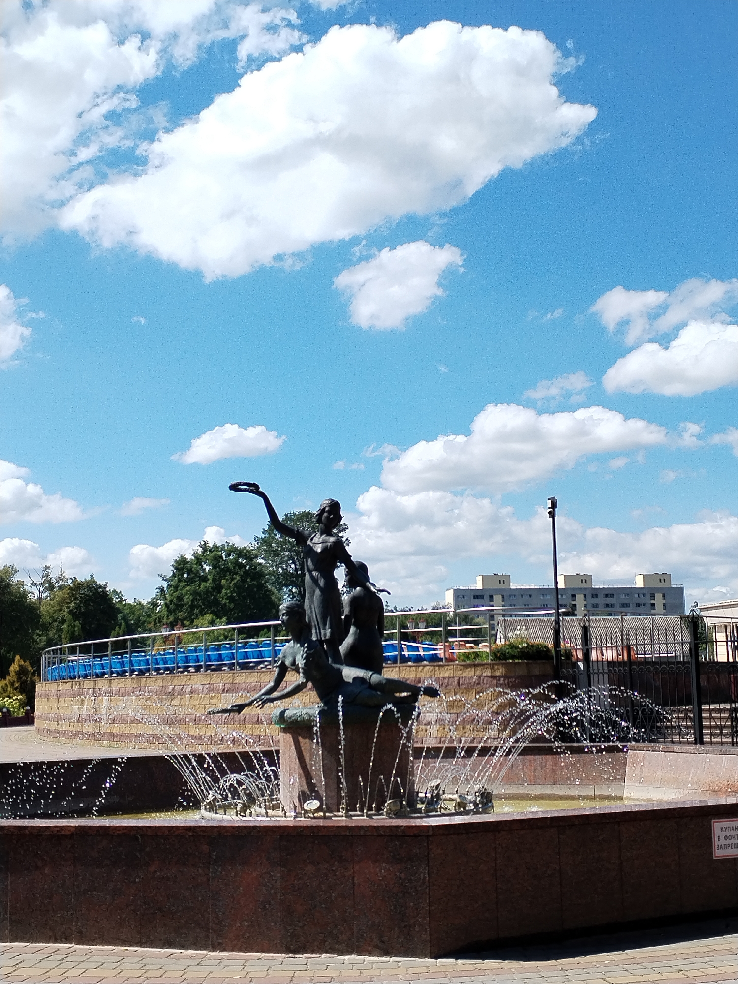 In the city of Unknown, Unknown, a bronze sculpture of three figures stands in a fountain under a bright blue sky with fluffy white clouds. The sculpture depicts a woman standing with her arms outstretched, holding a wreath or crown. Two other figures are partially visible, one kneeling and reaching forward, and another appearing to be seated. Water sprays from the fountain, creating a dynamic visual effect around the figures. Behind the fountain, a multi-tiered wall of rough-hewn stone is visible, topped with blue seating and a railing. In the background, lush green trees line the area, and beyond them, a modern, multi-story building with a light-colored facade and dark windows stands under the sky. A black lamppost is situated in front of the building. The foreground shows a paved walkway made of interlocking bricks in shades of red and beige. A small red sign with white text is affixed to the side of the fountain basin, though the text is too small to be legible.