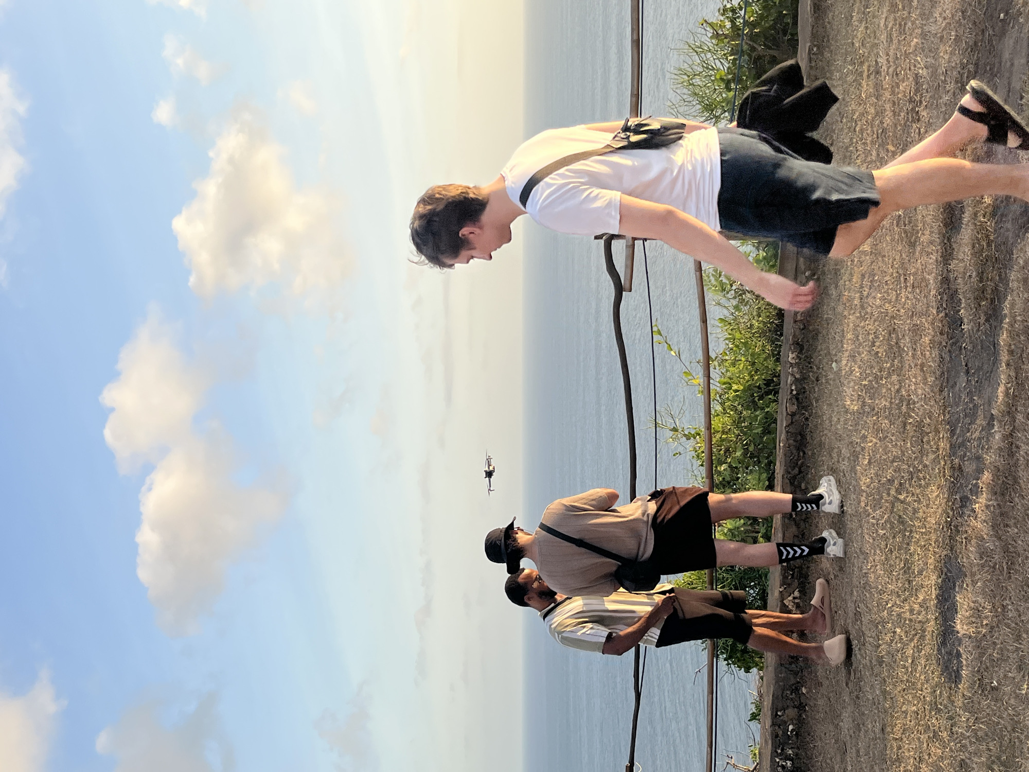Three individuals are standing on a grassy, elevated viewpoint overlooking the ocean. One person, wearing a white t-shirt and dark shorts, walks away from the camera to the right, carrying a dark bag slung across their chest and a dark jacket over their shoulder. Two other individuals stand together near a wooden railing, facing the ocean. The person on the left wears a striped short-sleeved shirt and dark pants, while the person on the right wears a beige short-sleeved shirt, a black hat, and dark shorts. A drone is visible flying in the sky between the groups of people and the ocean. The sky is blue with scattered white clouds, and the sun appears to be setting or rising, casting a warm light on the scene. The location is the coastal area of Kuta Selatan, Indonesia, known for its dramatic ocean views and popular tourist spots.
