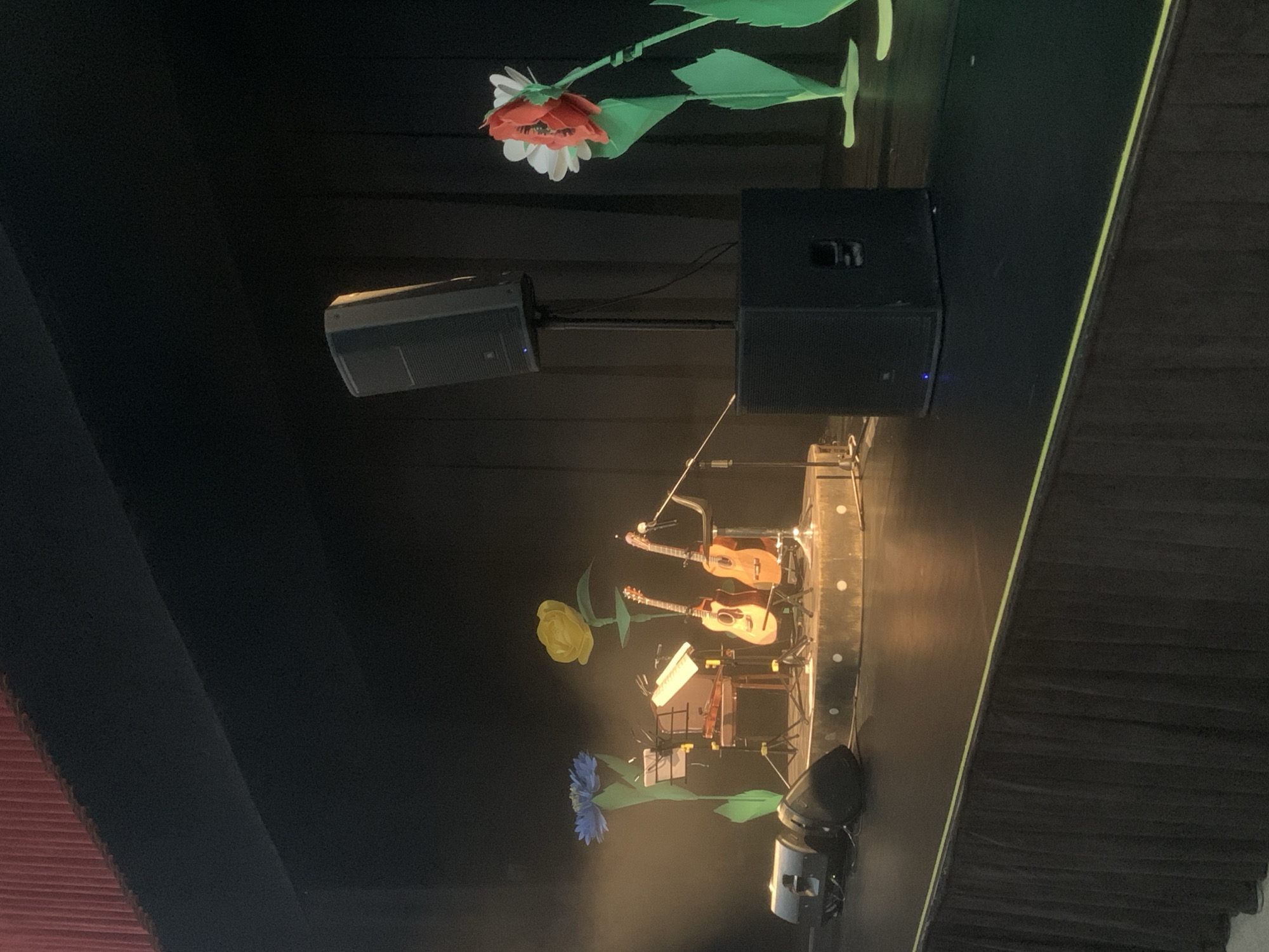 The image shows a stage setup, likely in Izhevsk, Russia. Two guitars are positioned on stands, flanked by musical equipment, including a keyboard and amplifiers. Decorative flowers, resembling oversized, whimsical artificial blooms, adorn the stage, one above the equipment, one to the left of the guitars, and one below. Stage lights are visible in the foreground. The stage has a dark backdrop and is likely set up for a performance. The curtain is to the left.